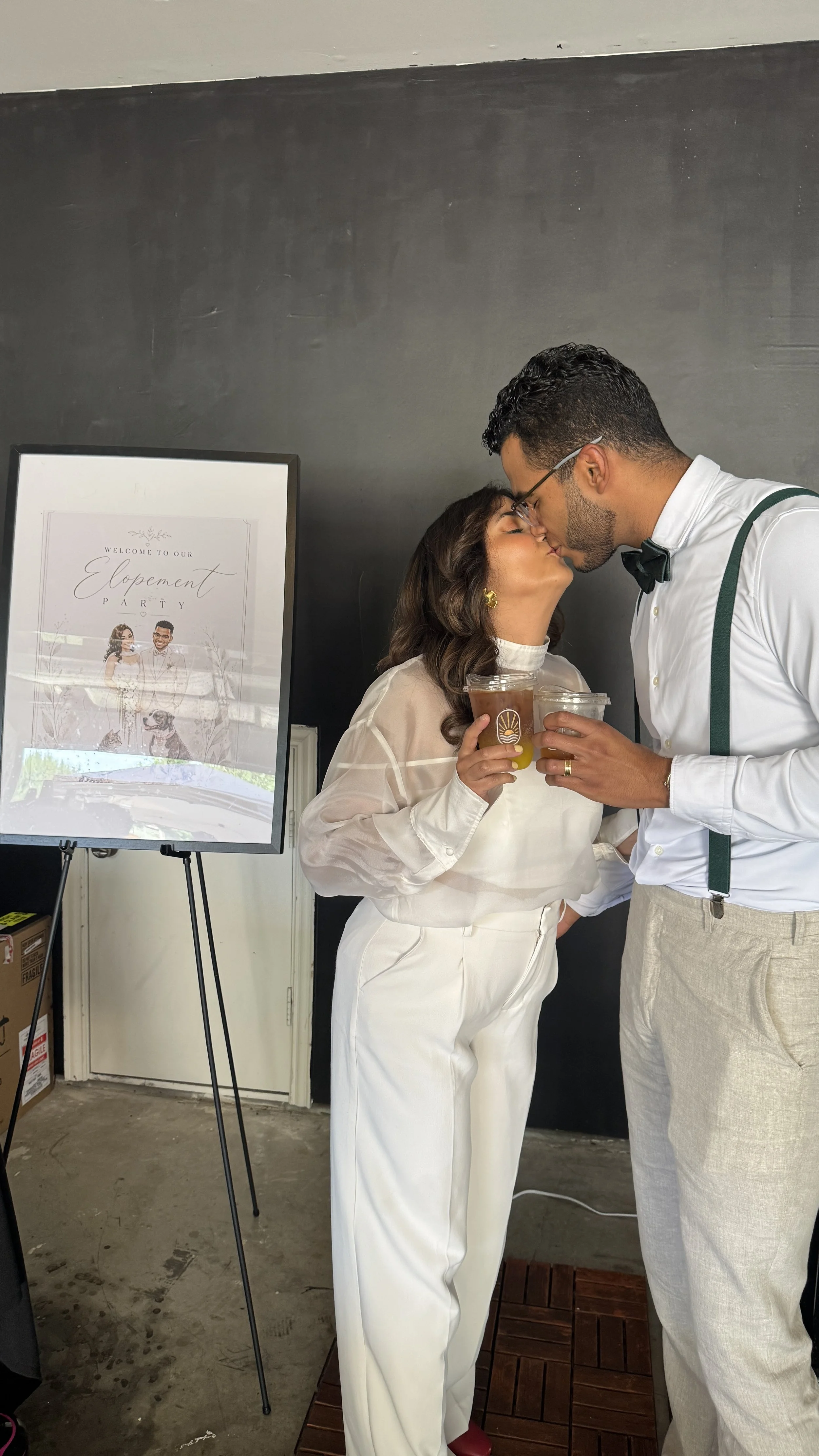 A man and woman are sharing a kiss at a celebration. The woman is holding two drinks and is dressed in white, while the man is wearing a white shirt with suspenders and a bow tie. A framed sign behind them reads 'Welcome to Our Engagement Party' with a picture of the couple and a dog.