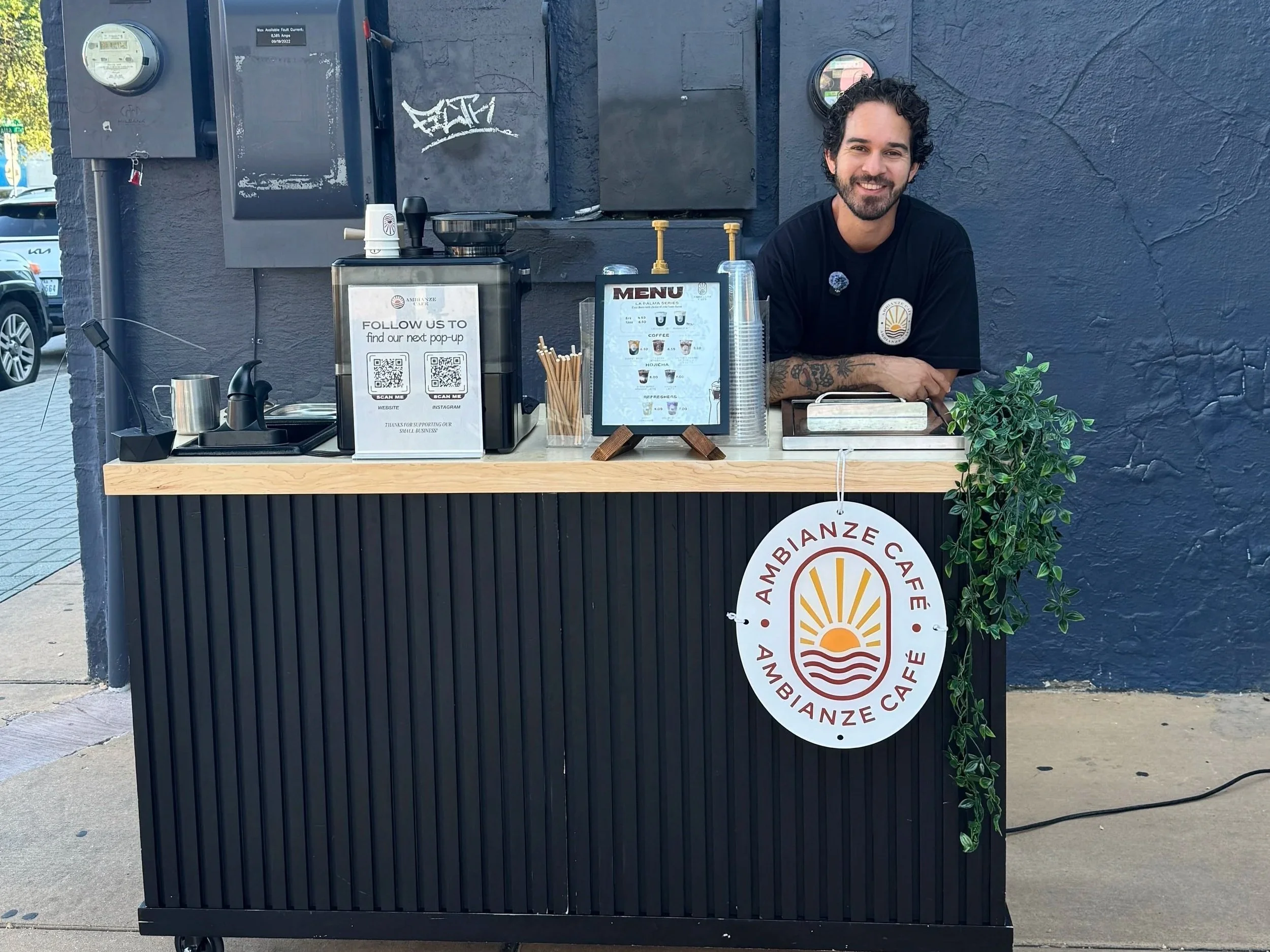 A smiling man with dark curly hair and a beard, standing behind a small outdoor café counter with a dark textured wall in the background. The counter has coffee-related signs, a plant on the right, and a menu displayed in a case, with a QR code for social media or website support.