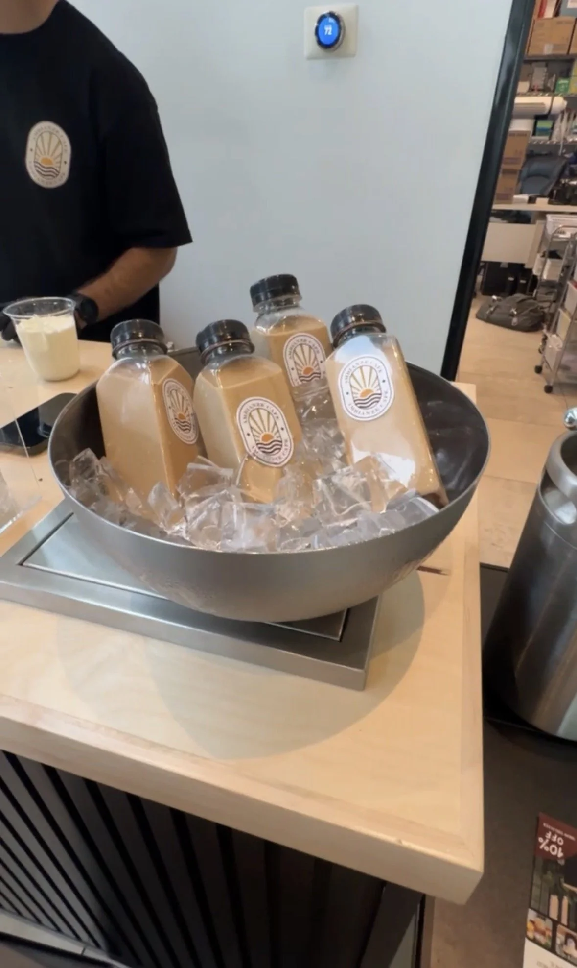 A bowl of bottled drinks with labels on ice. A person wearing a black shirt with a logo is partially visible in the background. A digital thermostat displays 72 degrees on a white wall.