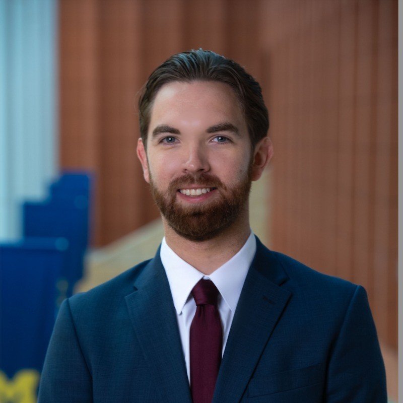 A young man in a navy suit, white shirt, and maroon tie, smiling, standing indoors with wooden and blue background.
