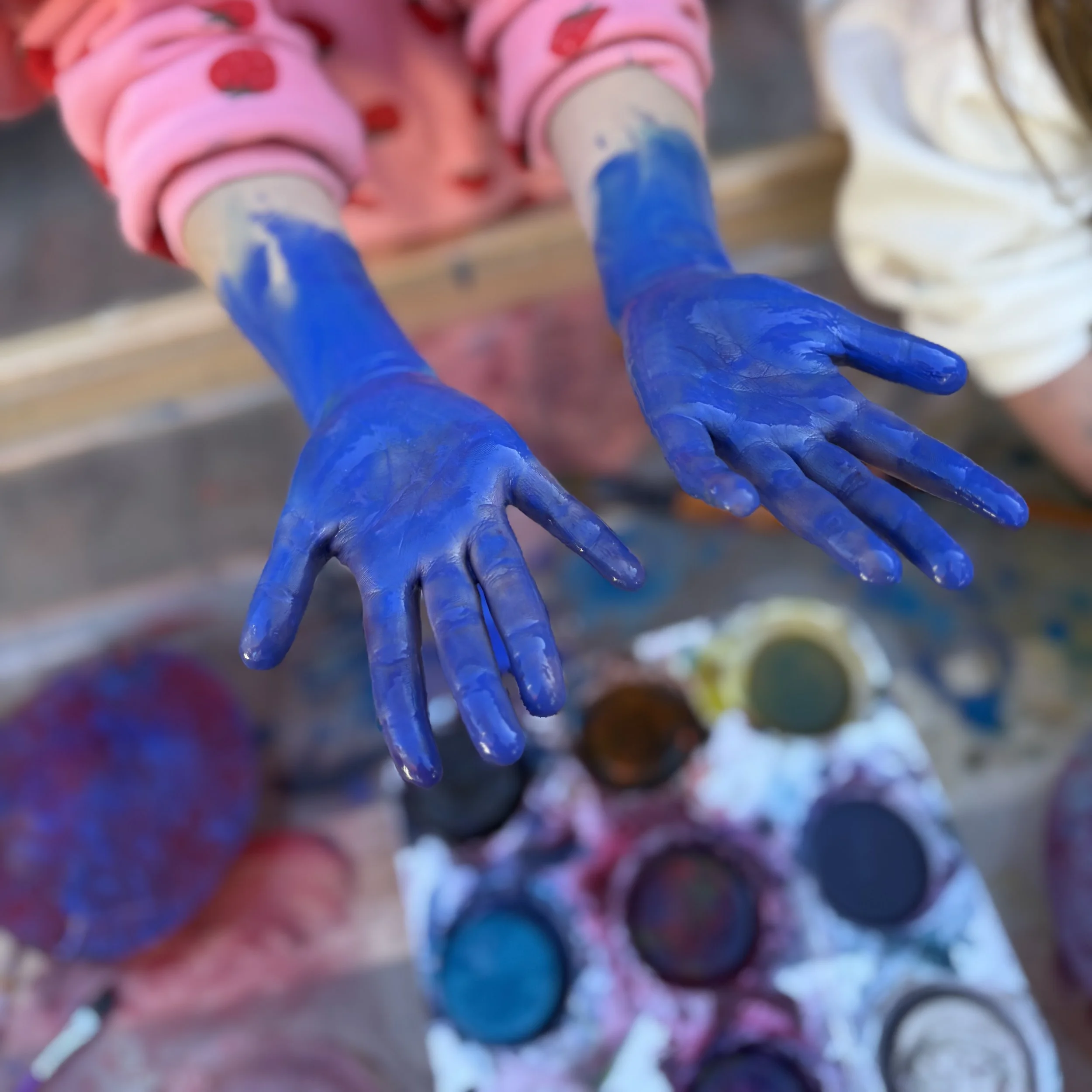 A child shows their hands that are covered in bright blue paint, with a pallet of paint colors in the background.