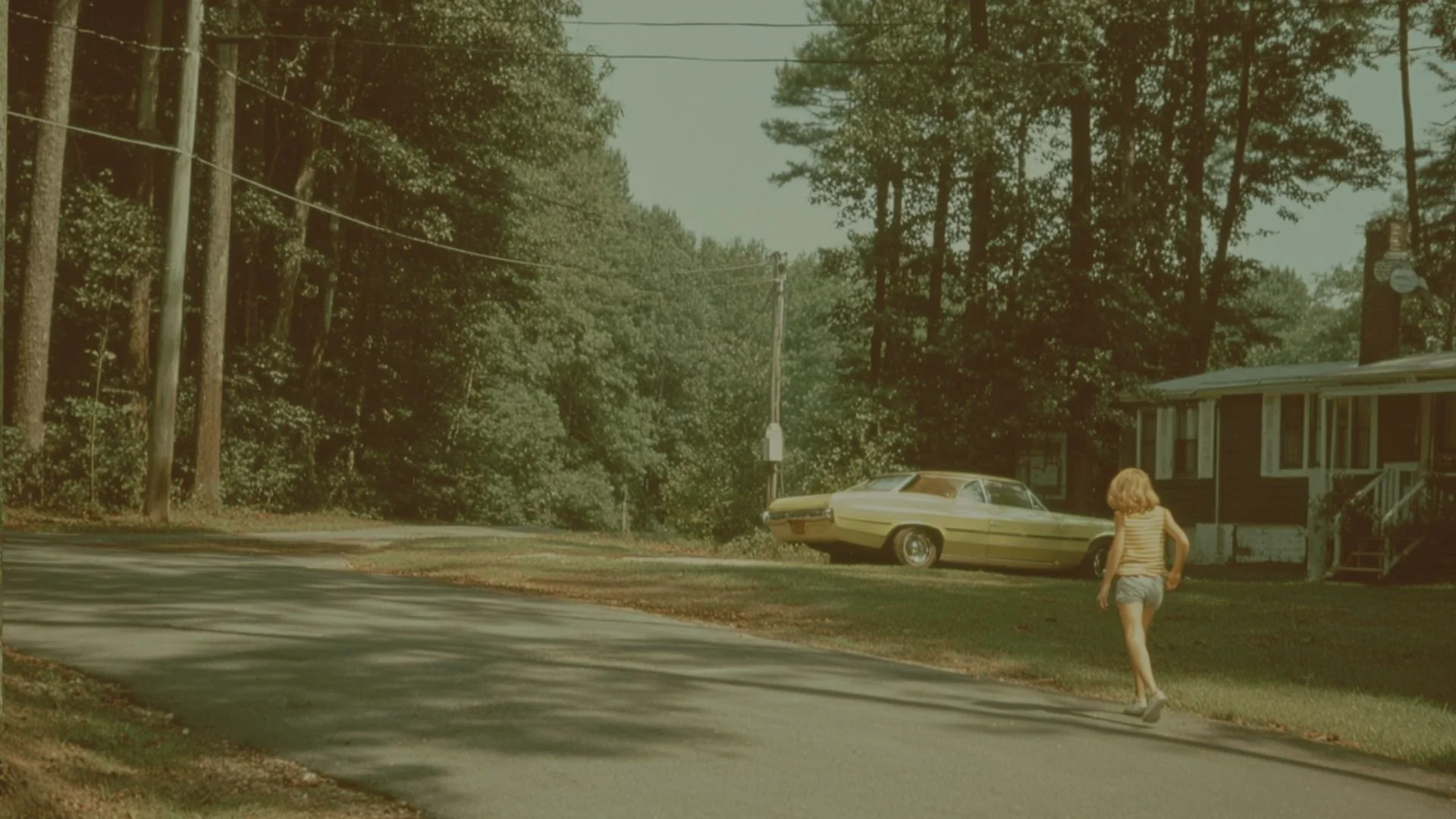 A girl with blonde hair wearing a striped tank top and shorts walking on a sidewalk near a yellow vintage car parked in front of a house surrounded by trees.