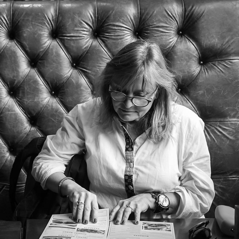 A woman with glasses and shoulder-length hair sits at a table, looking at a menu or pamphlet, with a tufted leather or faux leather headboard or wall behind her.