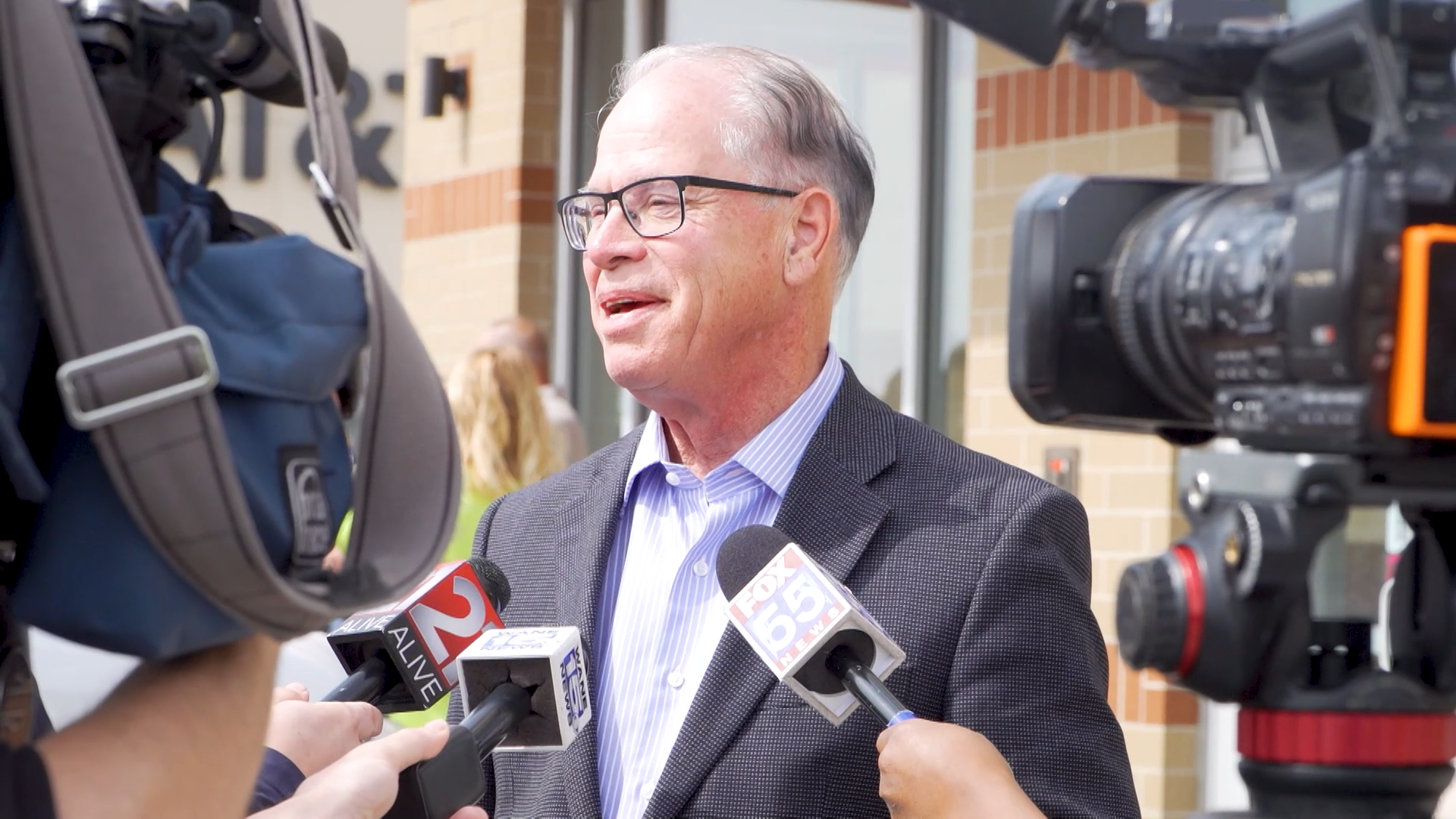 Man with glasses and a suit jacket speaking to reporters surrounded by microphones with news station logos, being filmed by a camera, outside a building with brick walls.