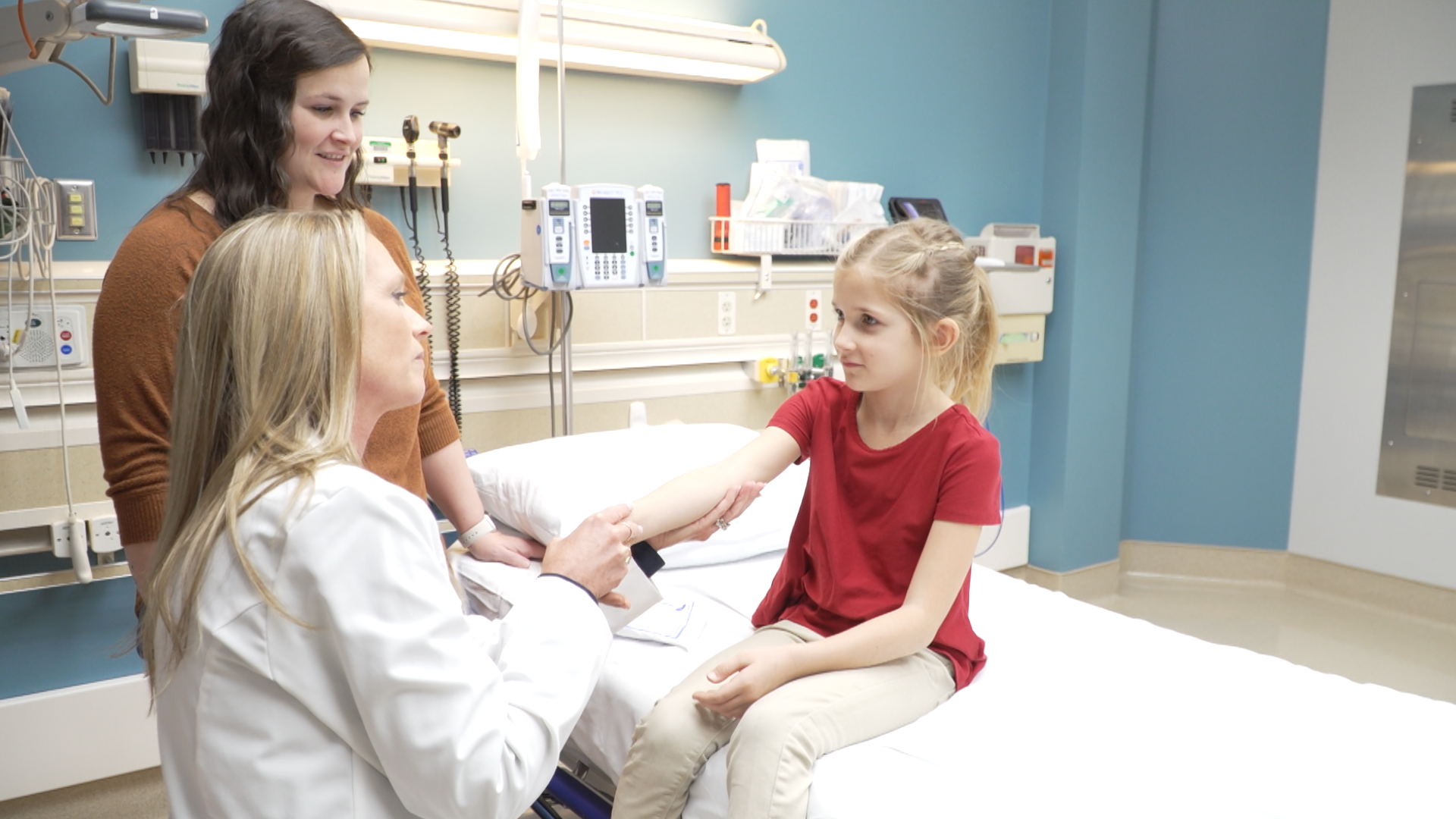 A young girl in a red shirt sitting on a hospital bed, being examined by a female doctor with long blonde hair. Two women, one with brown hair and one with blonde hair, are standing nearby in a hospital room with medical equipment and blue walls.