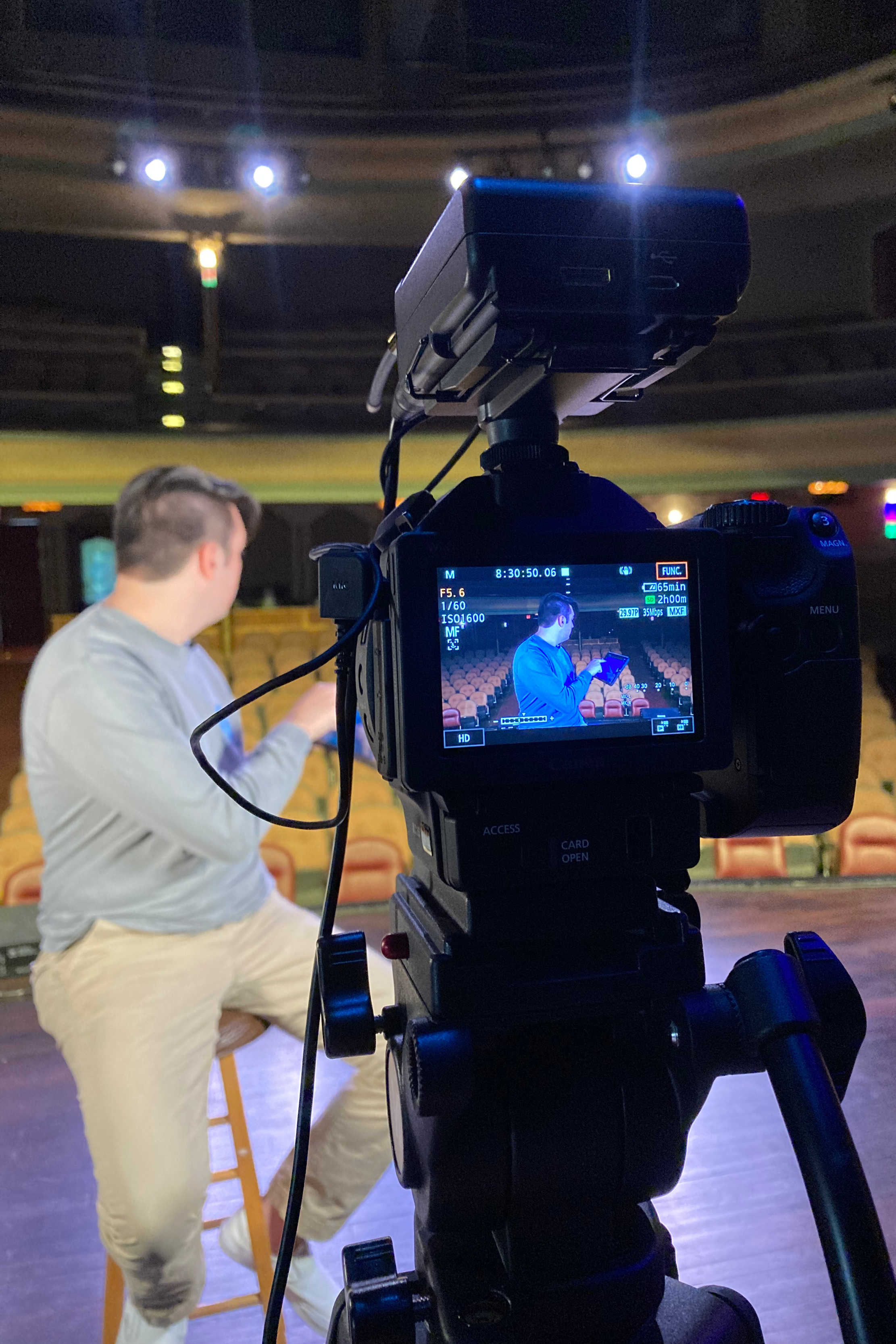 A man in a gray sweater and light-colored pants is sitting on a chair in an empty theater, holding a tablet, while a camera records him from in front of the stage.