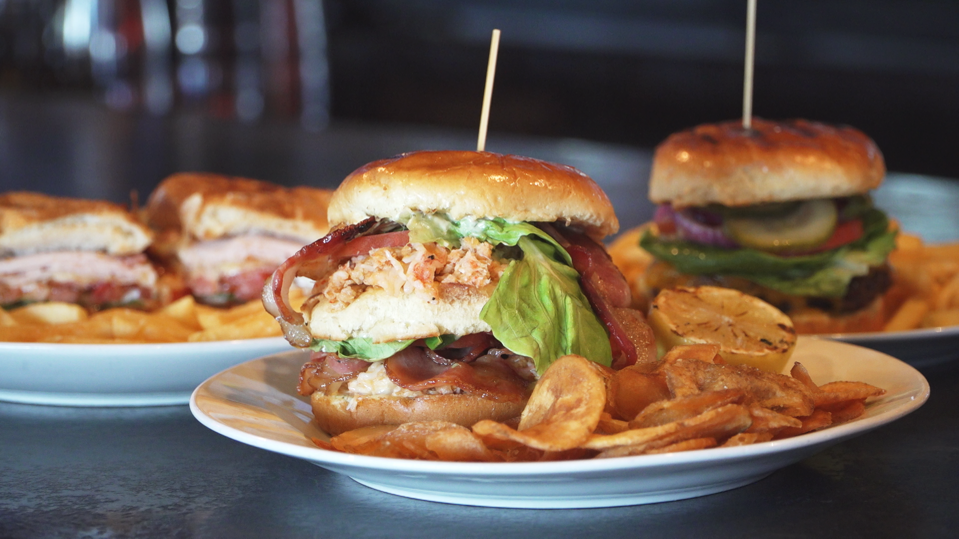 Plate with a bacon cheeseburger sandwich with lettuce, tomato, fried onion ring, and a side of potato chips. In the background, there are more sandwiches on plates and a dark countertop.