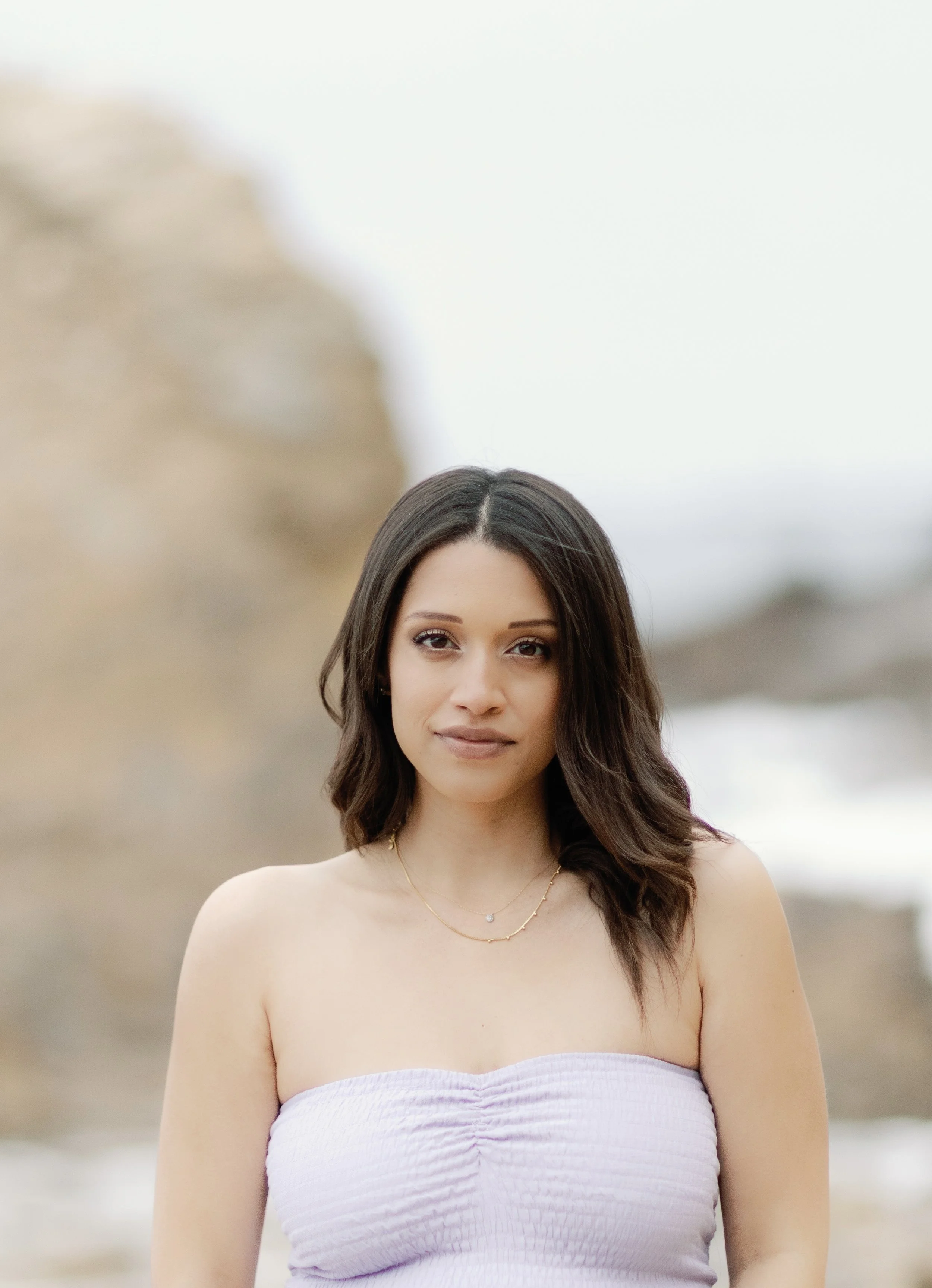 A young woman with long dark hair wearing a purple strapless dress and a delicate necklace standing outdoors near a body of water and a grassy area.
