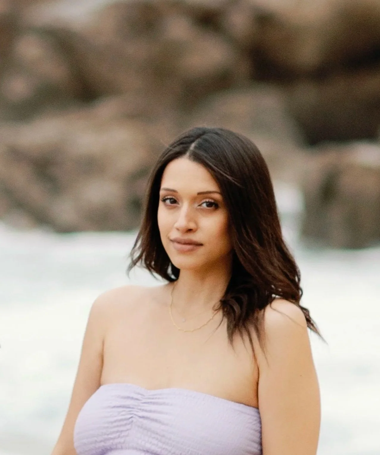 A young woman with dark brown hair wearing a strapless white top standing outdoors near water with rocks in the background.
