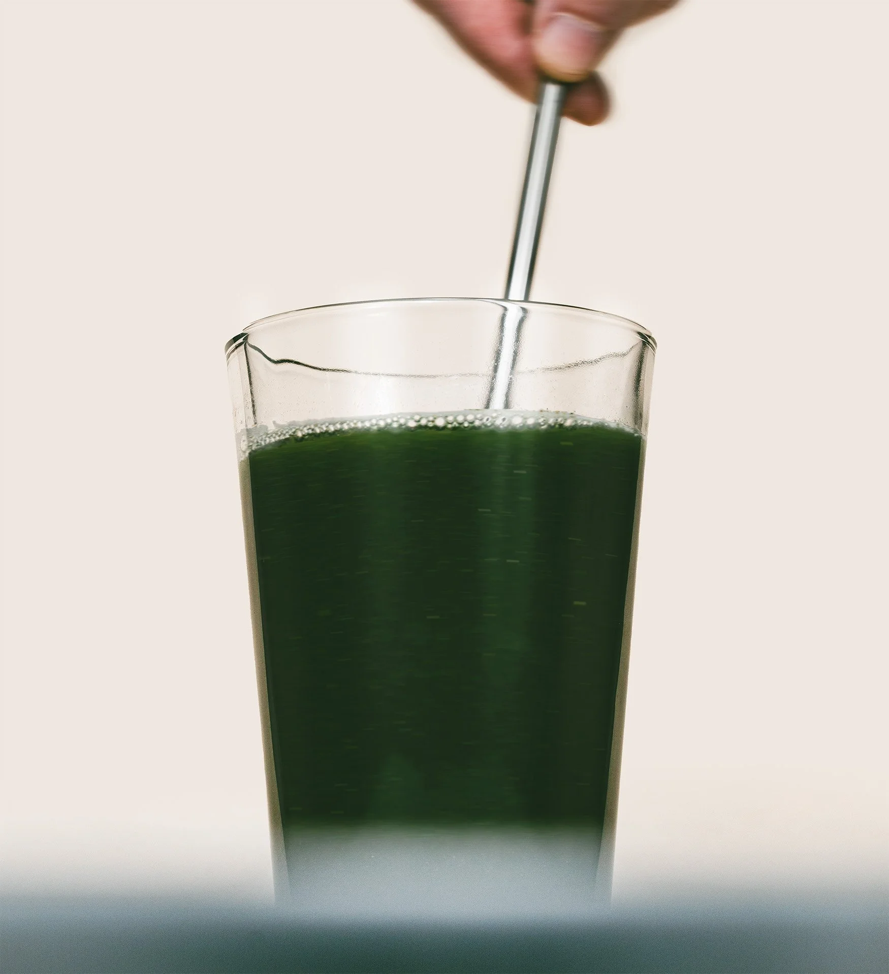 A glass of green juice being stirred with a metal straw held by a person's hand.