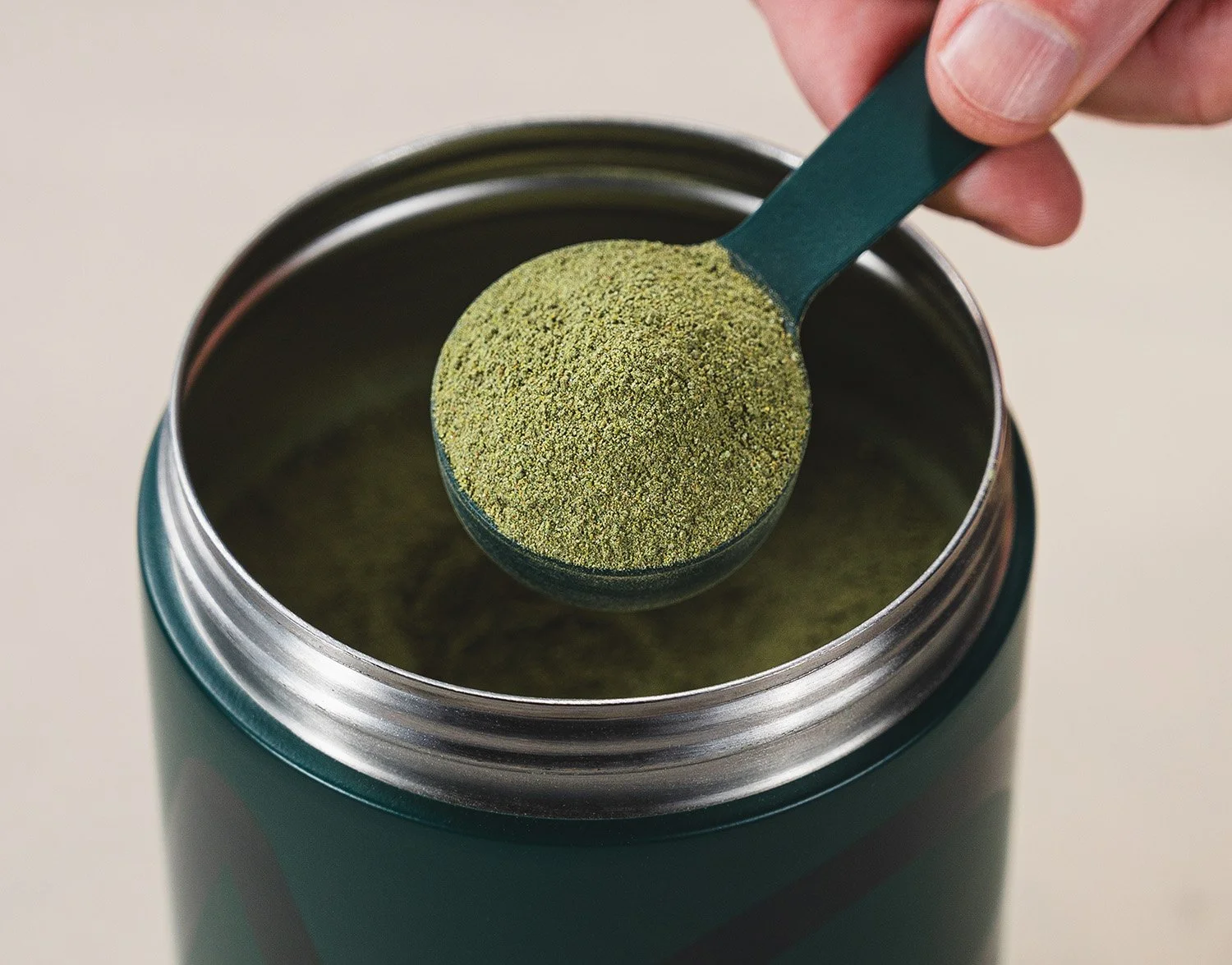 Close-up of a hand using a green scoop to transfer green powdered supplement from a teal container.