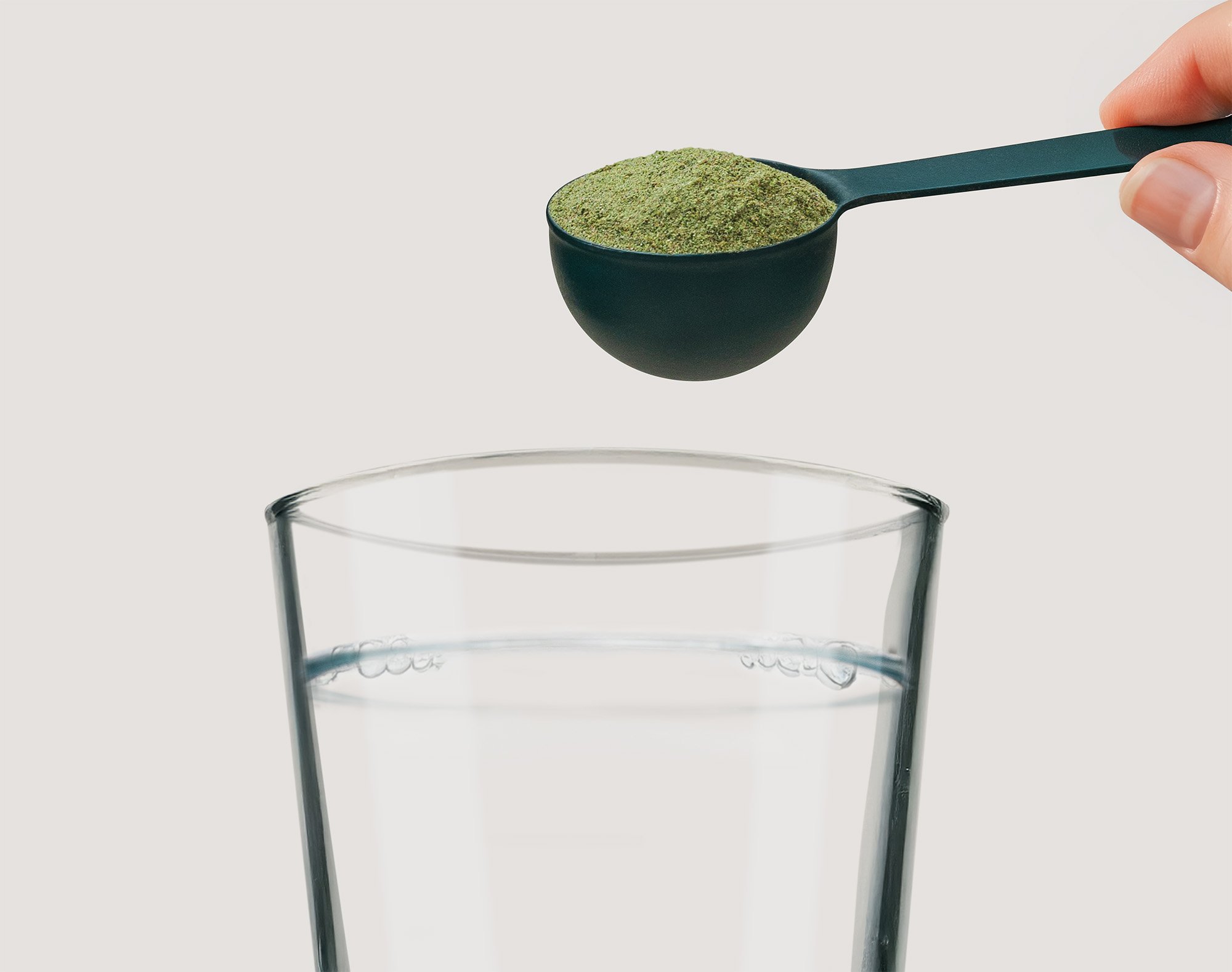 A hand holding a black measuring spoon filled with green powder above a glass of water.