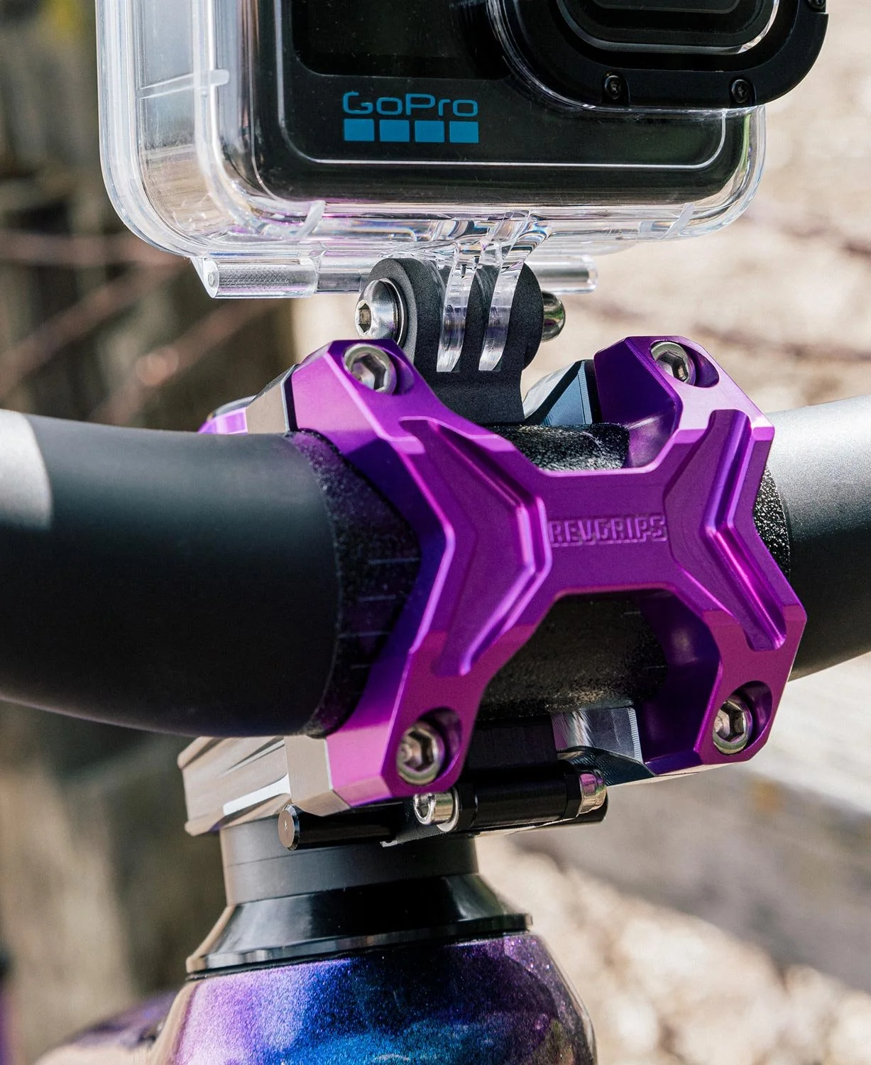 Close-up of a purple bicycle handlebar with a Revespikes stem, a GoPro camera mounted in a clear waterproof case, all outdoors.
