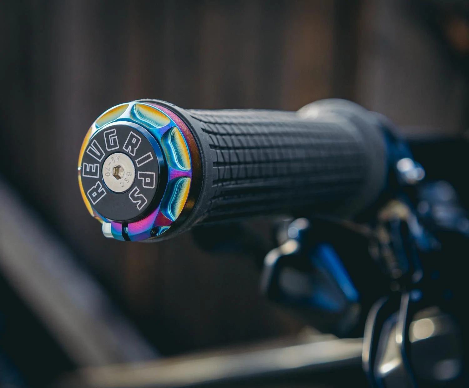 Close-up of a black handlebar grip on a motorcycle, showing the end cap with a colorful, iridescent finish and branding that reads "REVGRIPS SS USA".