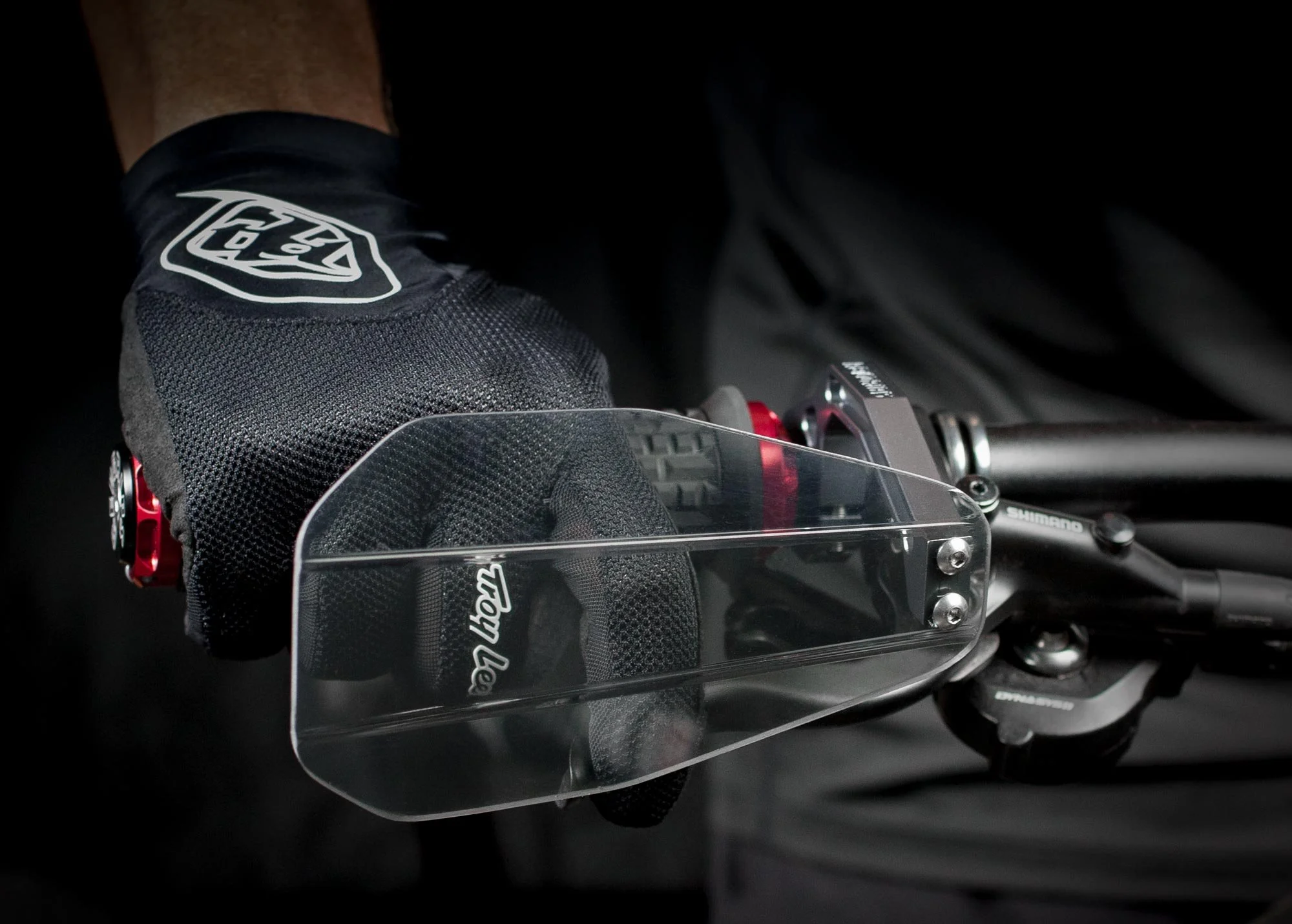 Close-up of a cyclist's hand wearing a black glove with a white logo, gripping the handlebar of a mountain bike with a clear windscreen attached to the handlebar.