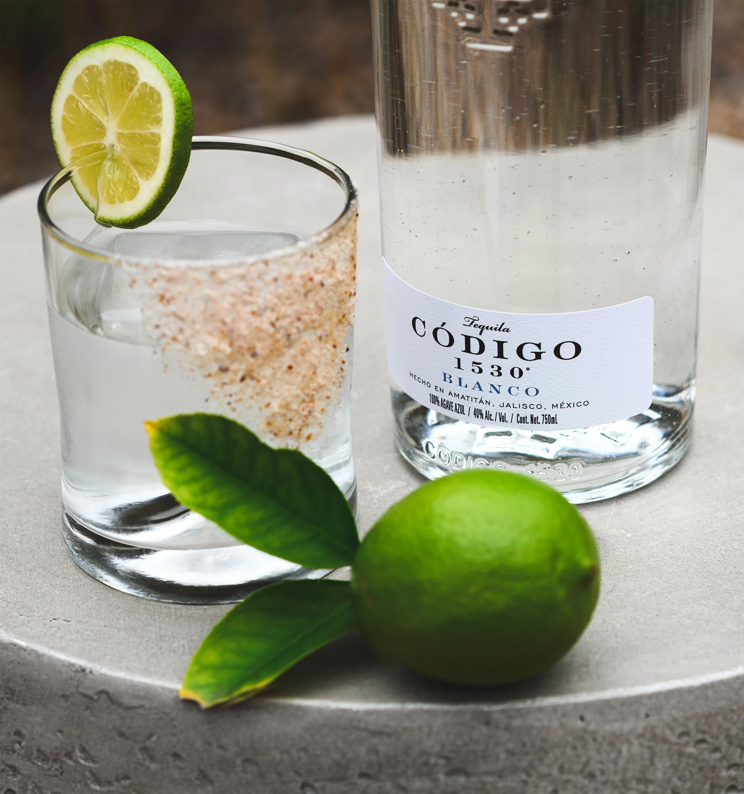 A tequila bottle labeled 'Código Blanco' next to a glass with salt and a lime slice, with green lime and lime leaves in the foreground.