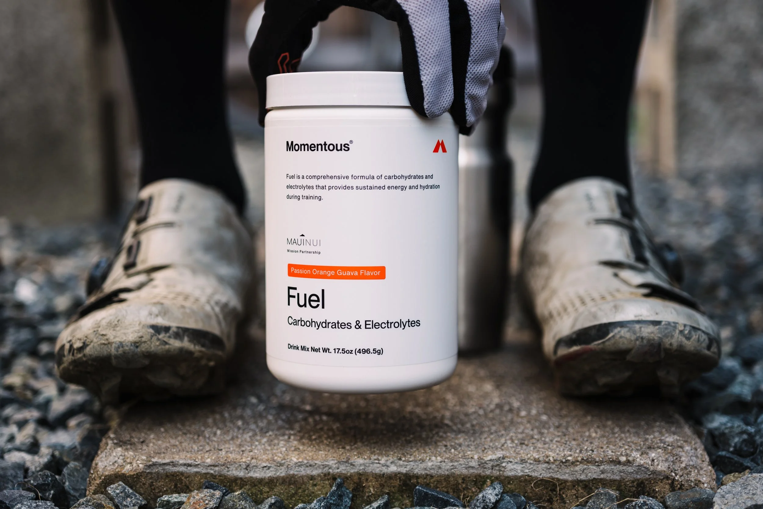 A person wearing camouflage shoes and black socks sitting on rocky ground outdoors, holding a white container labeled 'Momentous Fuel,' which contains carbohydrates and electrolytes for energy during training.