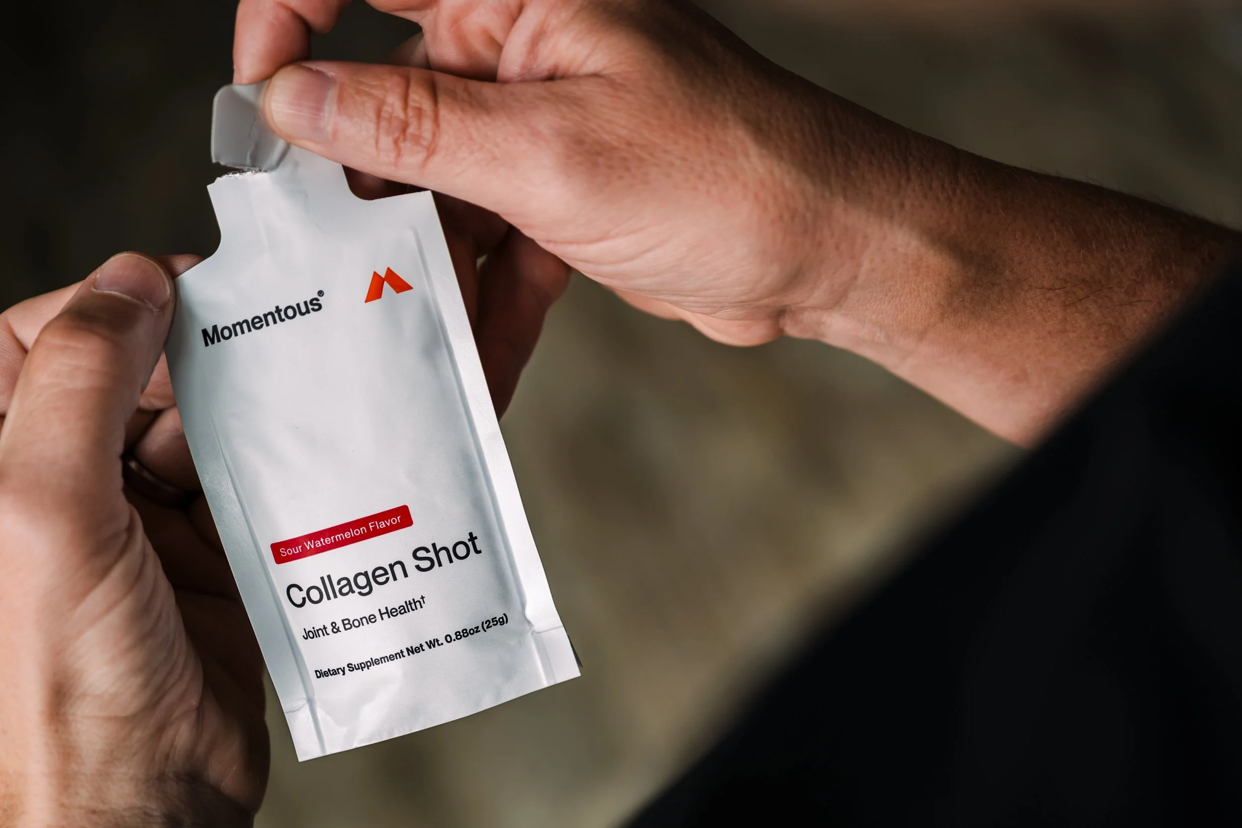 Close-up of a person holding a packet labeled 'Momentous' that contains a supplement called 'Hagen Shot' with sour watermelon flavor, for joint and bone health.