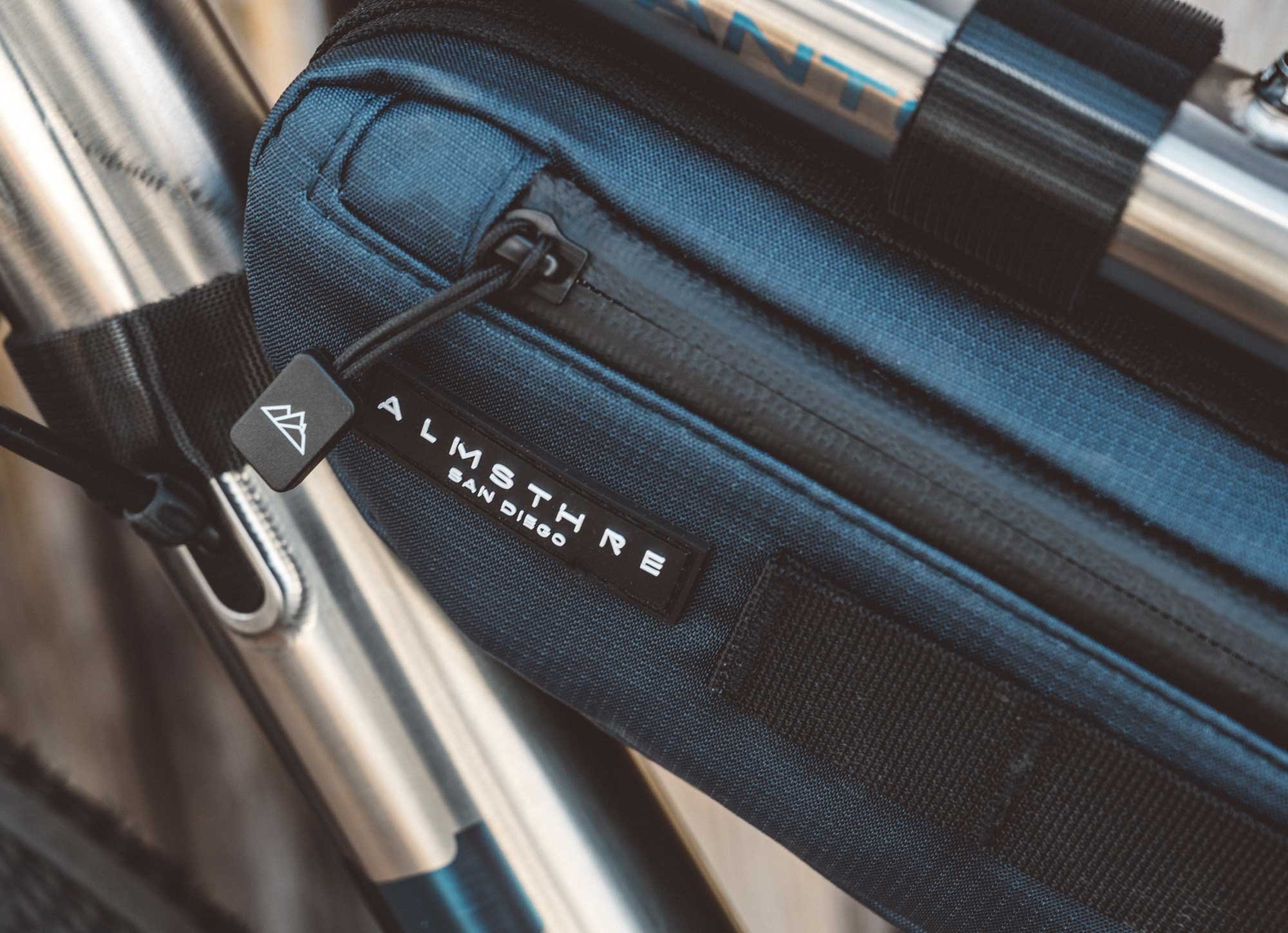 Close-up of a black and blue bag with a zipper, attached to a luggage handle, with a tag that reads 'ALMSTORE SAN DIEGO'.