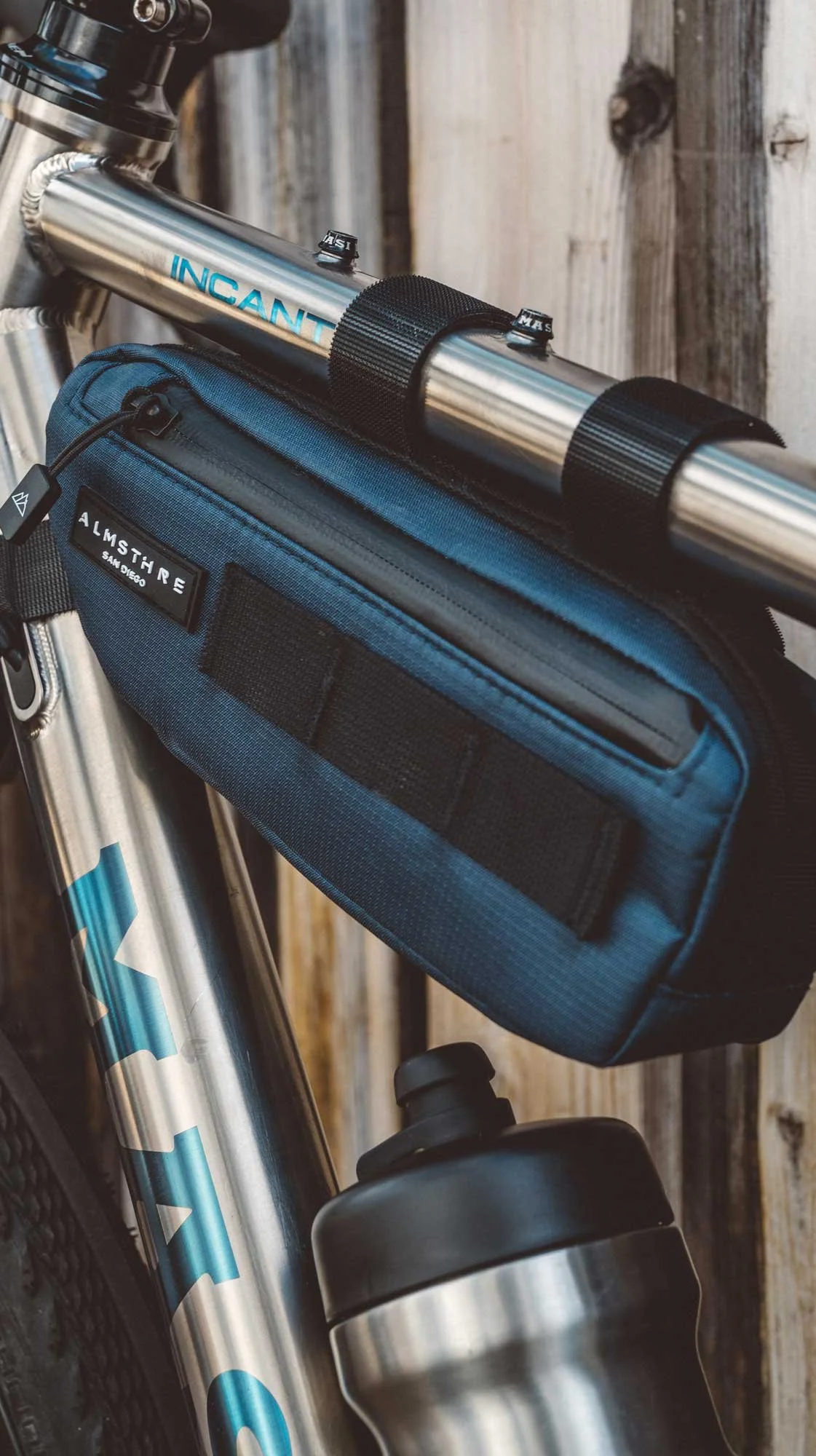 Close-up of a bicycle's front part including a black water bottle, a blue handlebar bag labeled Alma and a silver frame, with a wooden fence in the background.
