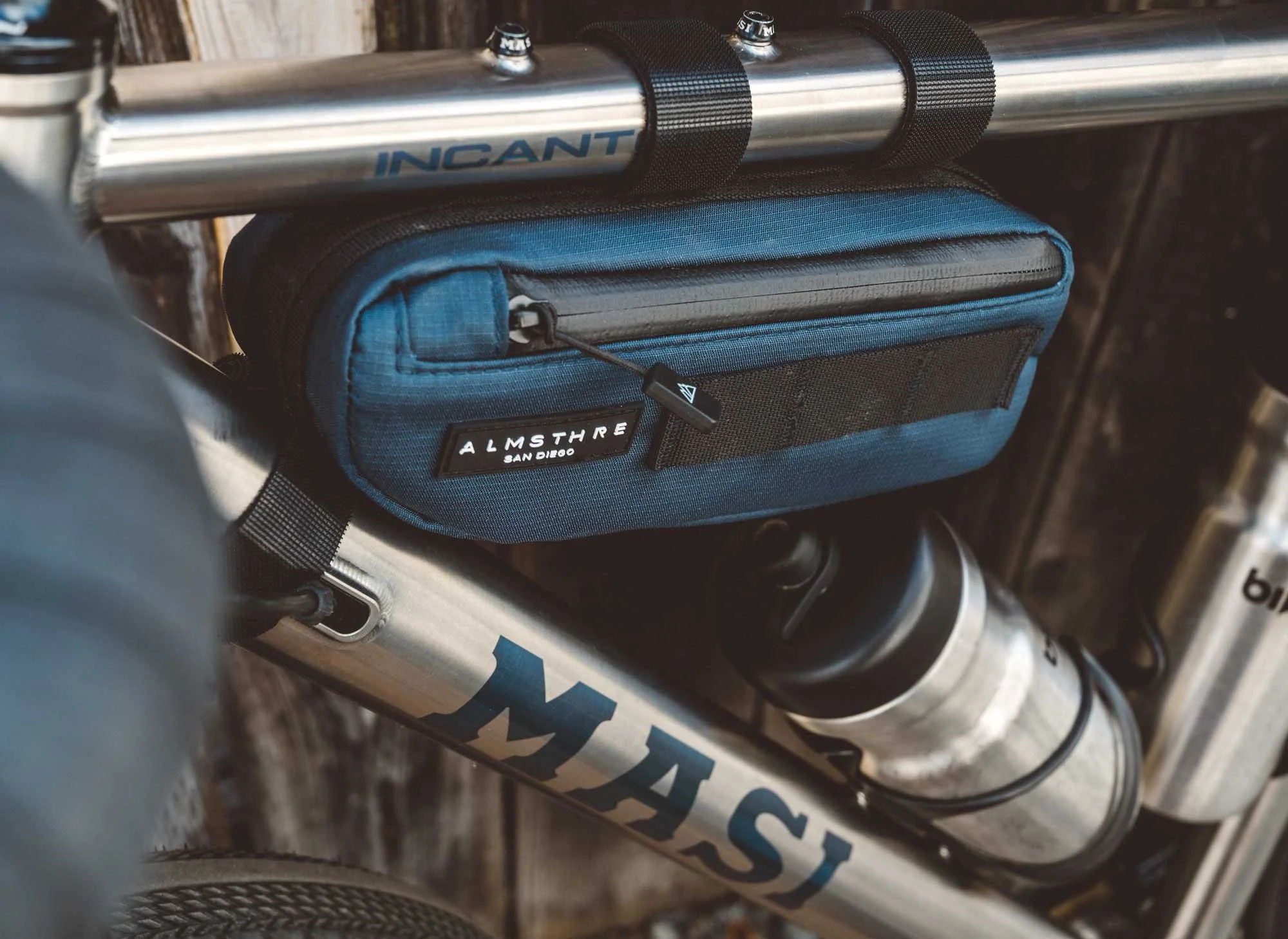 Close-up of a bicycle handlebar with a blue Almsothe San Diego pouch attached and a stainless steel water bottle mounted on the frame.