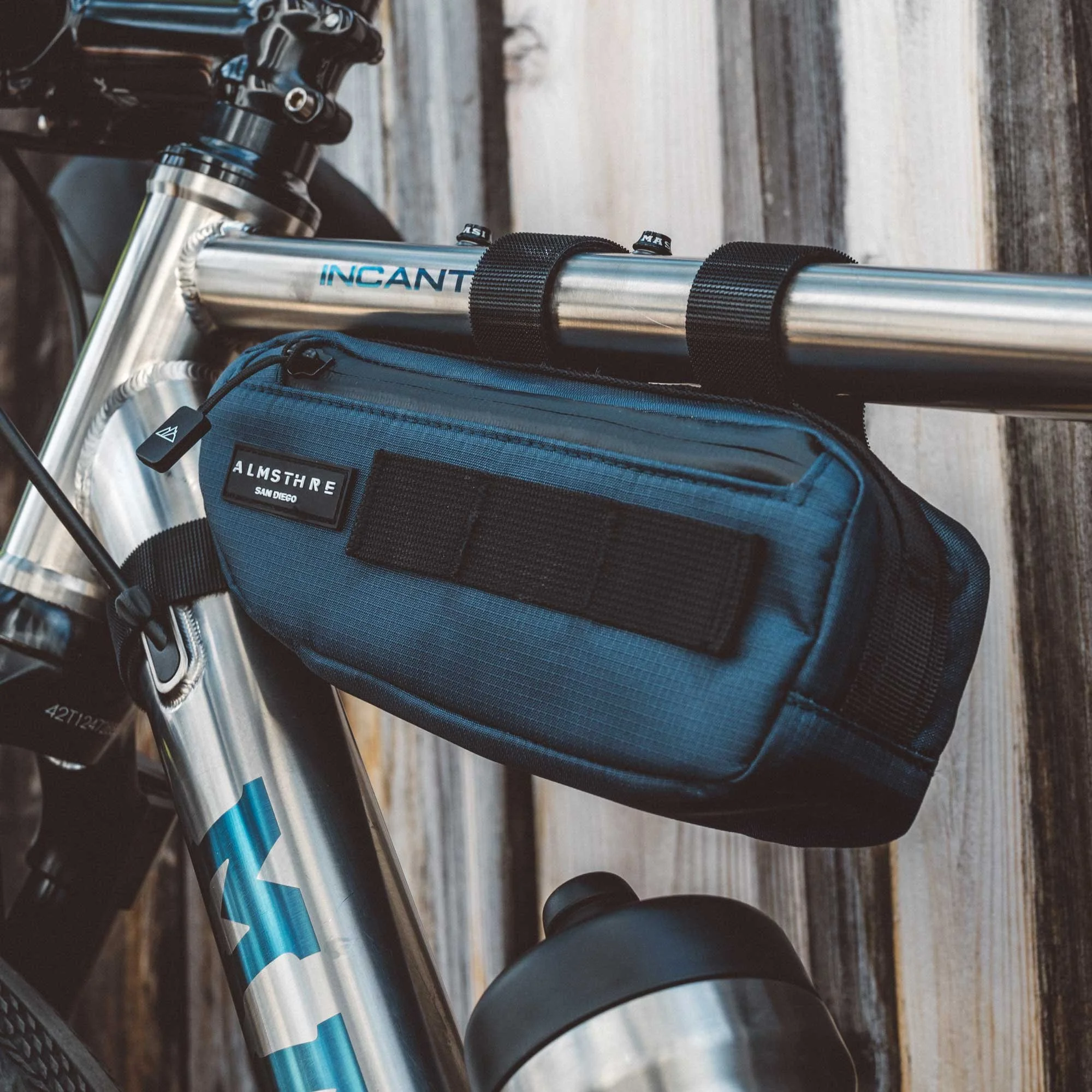 Close-up of a silver mountain bike with a blue frame, mounted with a black and blue ALMSHARE saddlebag attached to the top tube, against a wooden background.