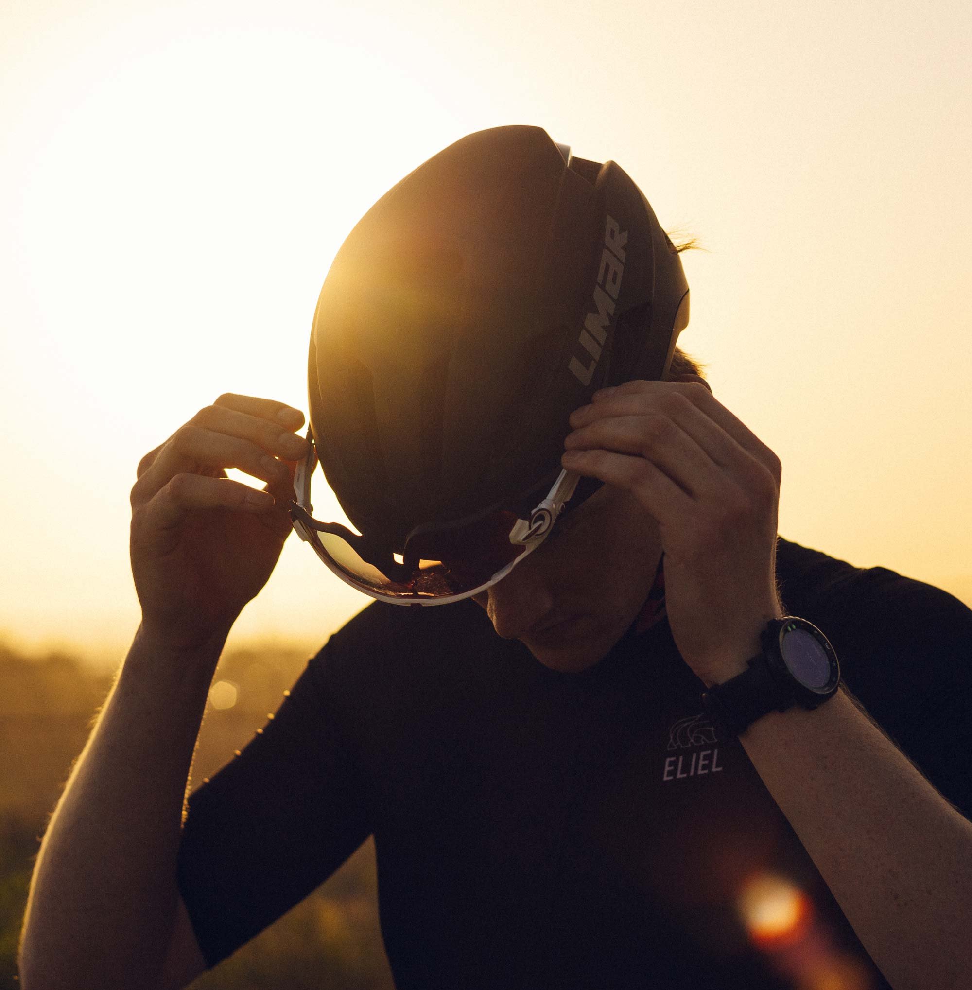 Person holding a motorcycle helmet with a sunset or sunrise background.