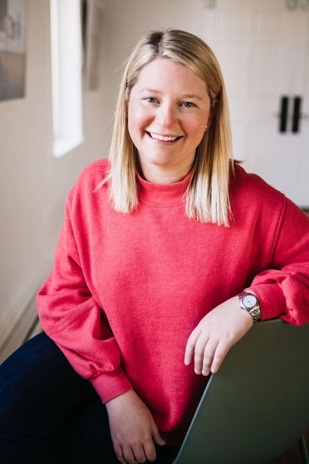 A woman with blonde hair wearing a red sweatshirt and a watch, smiling and leaning on a laptop in a bright room with natural light.