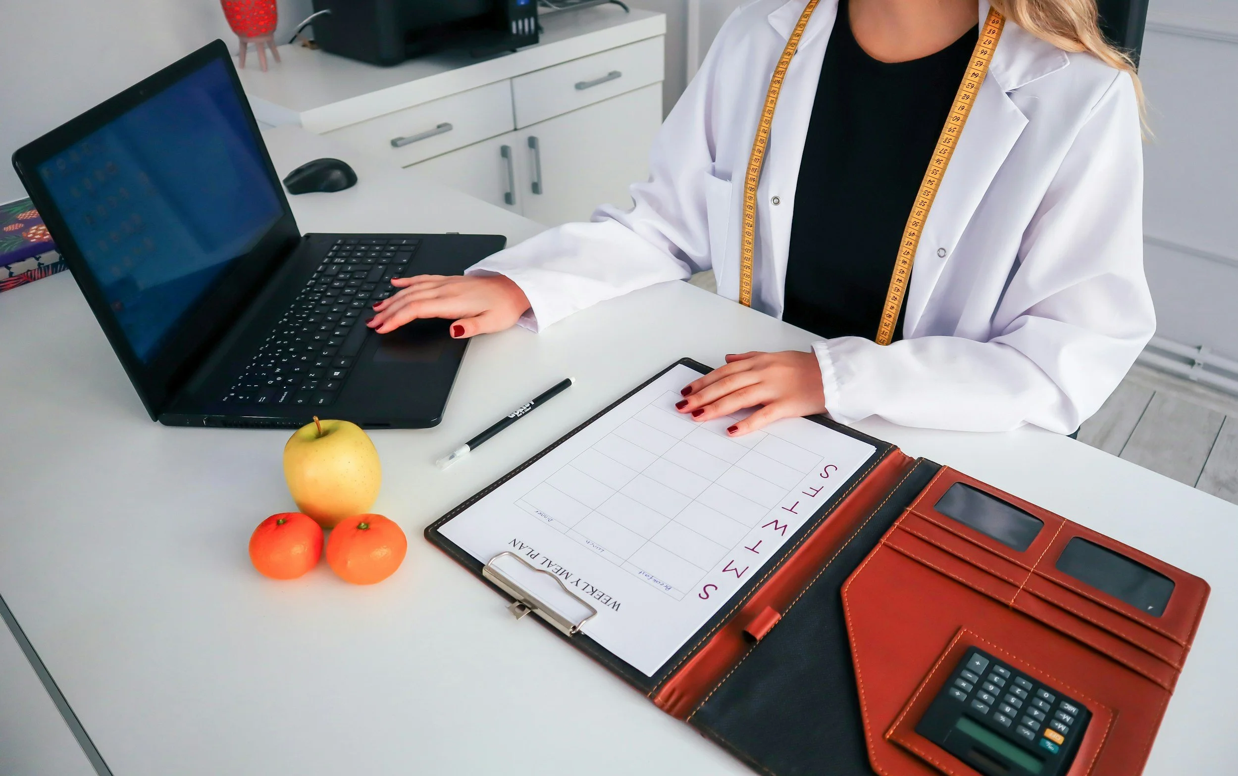 A person in a white coat with a tape measure around their neck sitting at a white desk with a laptop, a black calculator, a clipboard, and a notebook with a weekly meal plan. There are also three apples and two oranges on the desk.