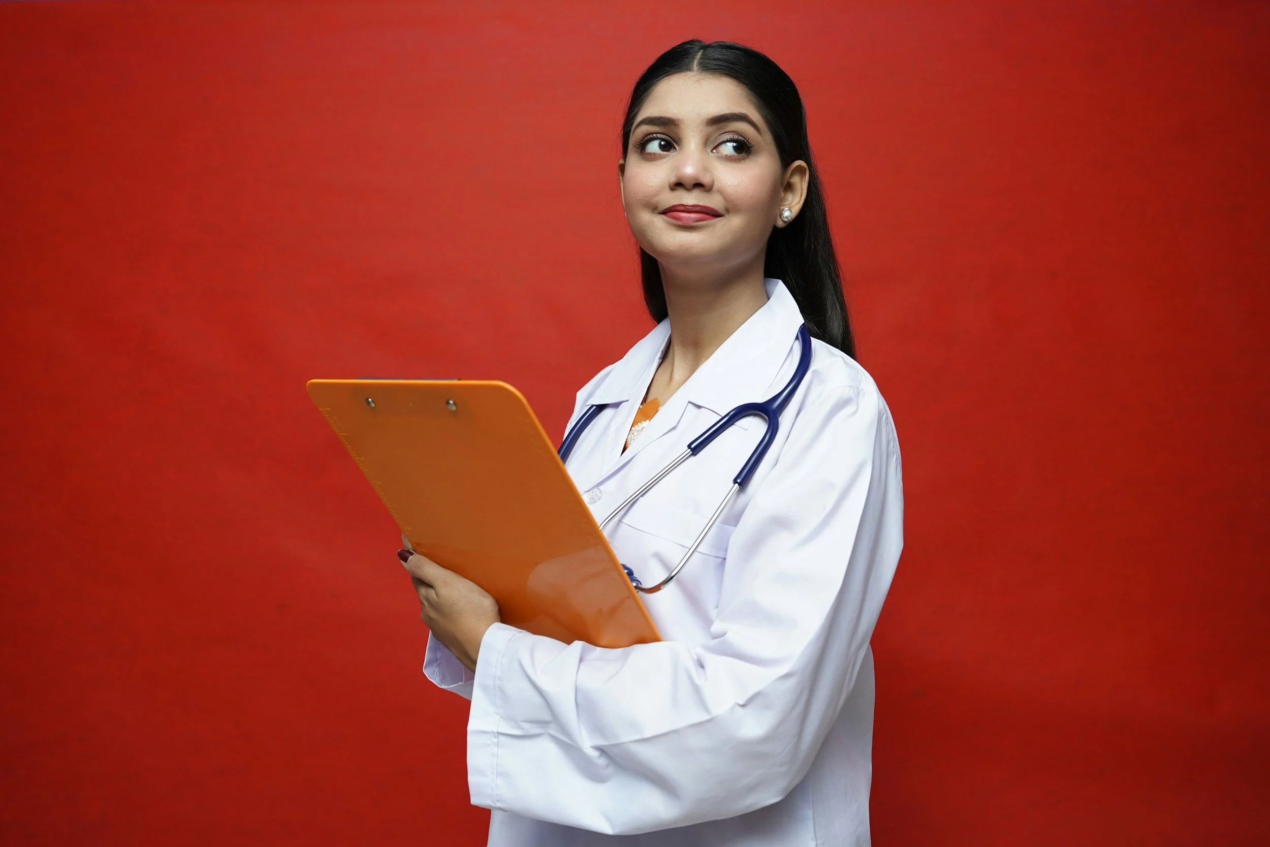A young female doctor with dark hair, wearing a white coat and a stethoscope around her neck, holding an orange clipboard, standing against a red background, looking to her right with a slight smile.