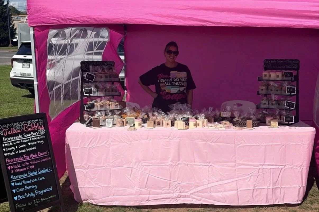 A woman stands behind a pink table at a soap stand, smiling, with soap products displayed on the table and on multi-tiered stands. A chalkboard sign listing soap flavors is positioned to the left of the table.