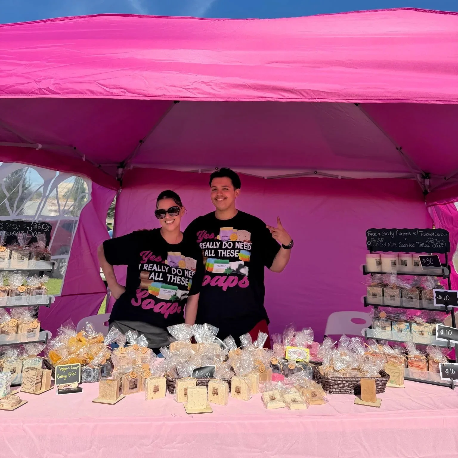 Two smiling people standing behind a table at a market stall selling handmade soap, with pink canopy. They wear matching black T-shirts with colorful text and images, and there are various soap products in front of them labeled with prices.