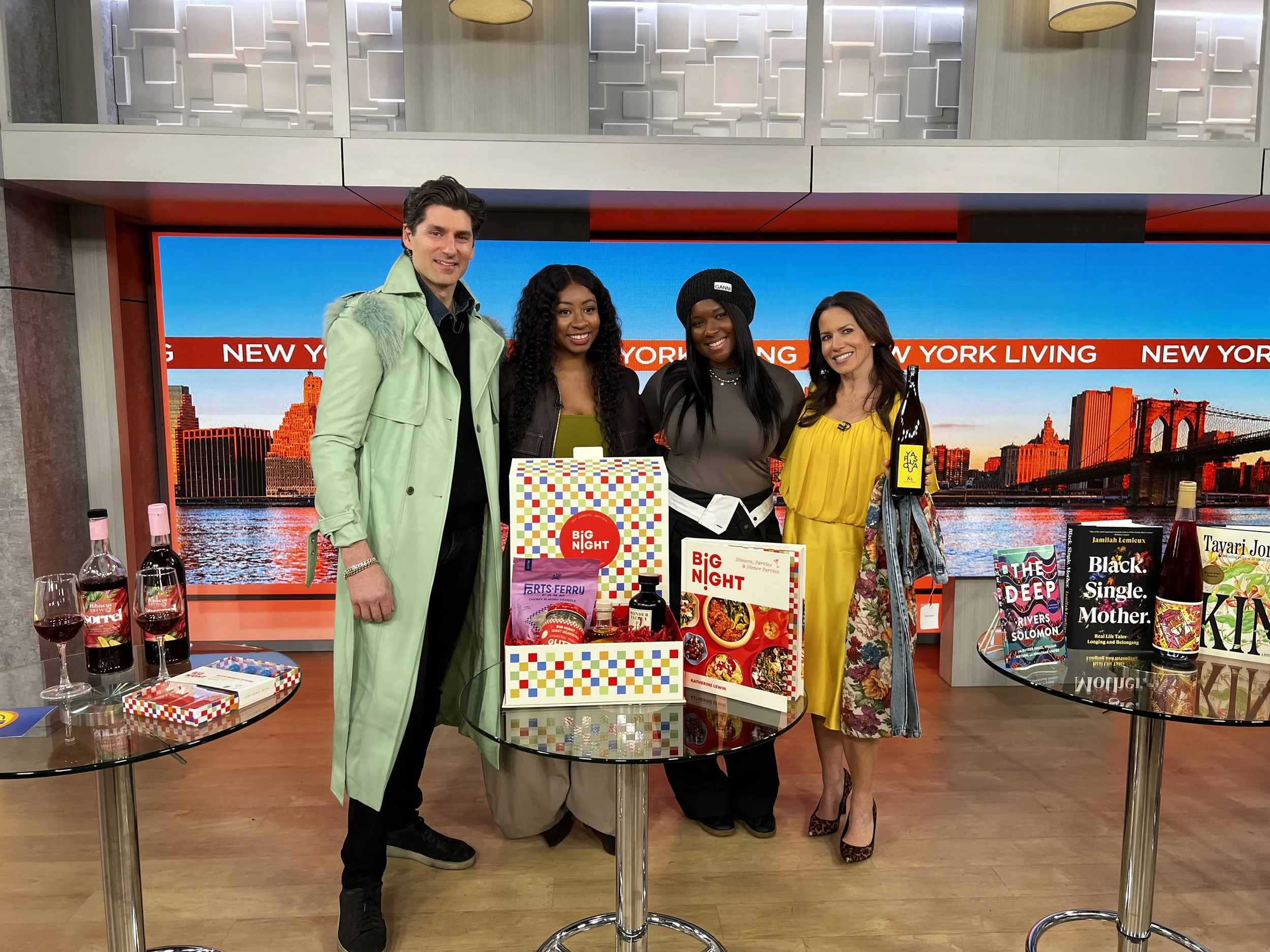 Four people standing behind a display table with books and wine bottles, in front of a large digital screen showing the New York City skyline and the Brooklyn Bridge.