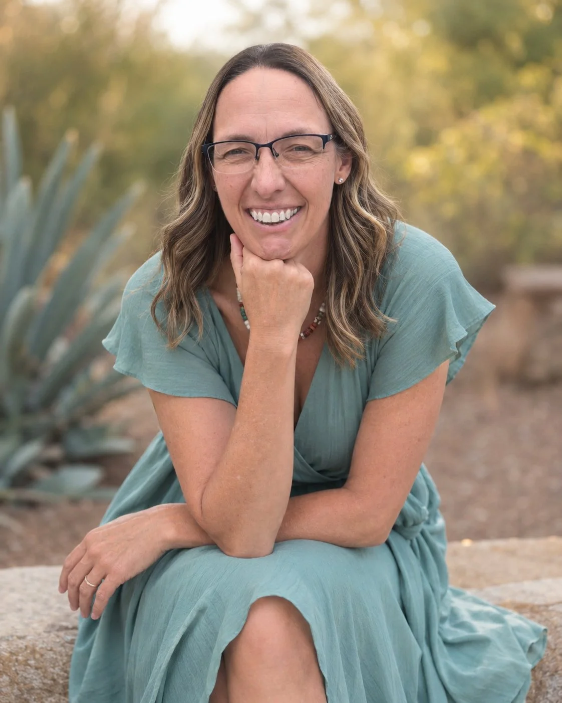 A woman with glasses and long wavy hair, sitting outdoors on a rock, smiling, wearing a blue dress, with a blurred background of trees and plants.