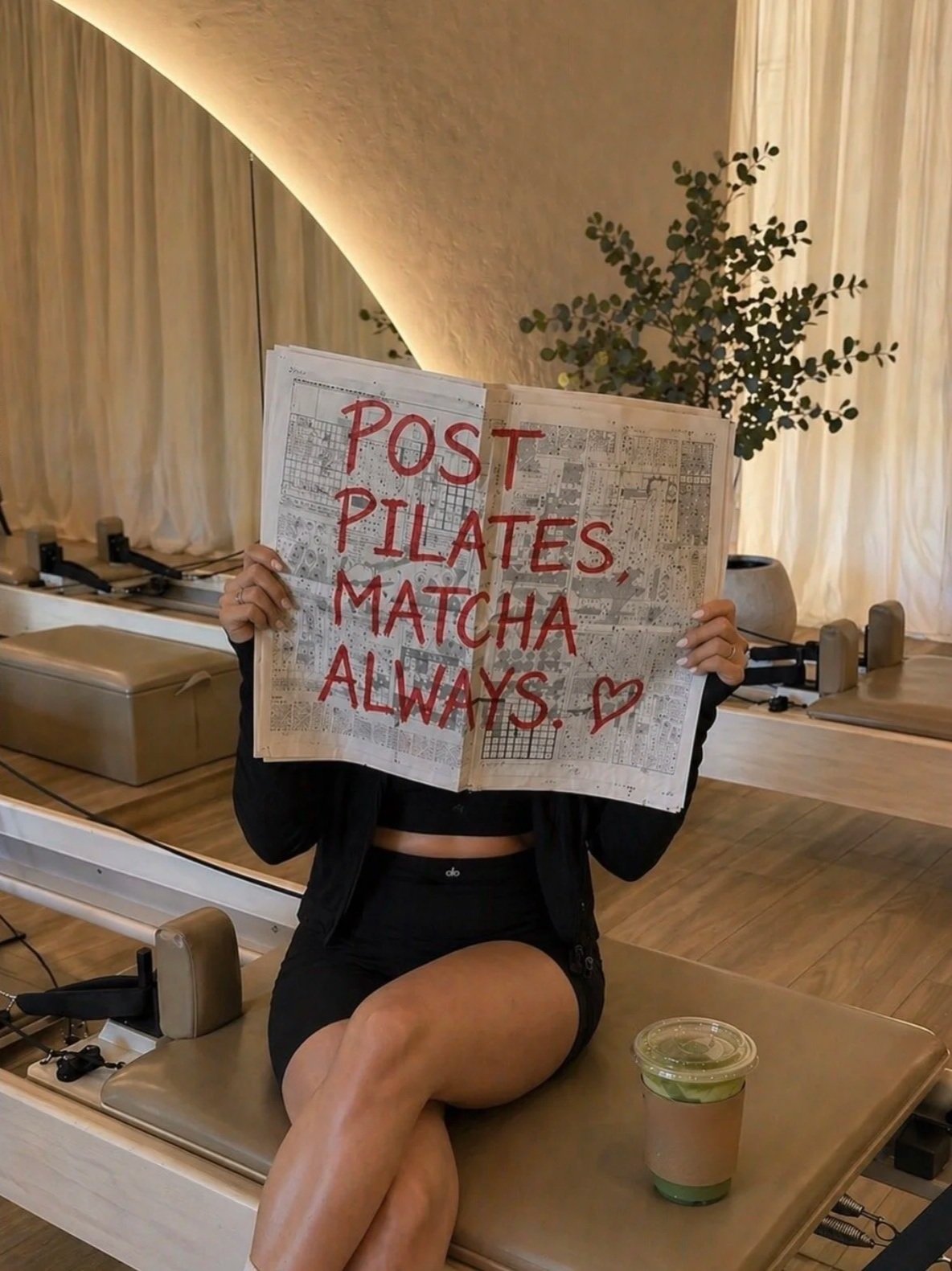 Person sitting on a massage table holding a newspaper with a sign in red letters that says "POST PILATES, MATCHA ALWAYS" with a heart. The person is wearing black workout clothes and has crossed legs. There is a matcha drink on the table in front of 