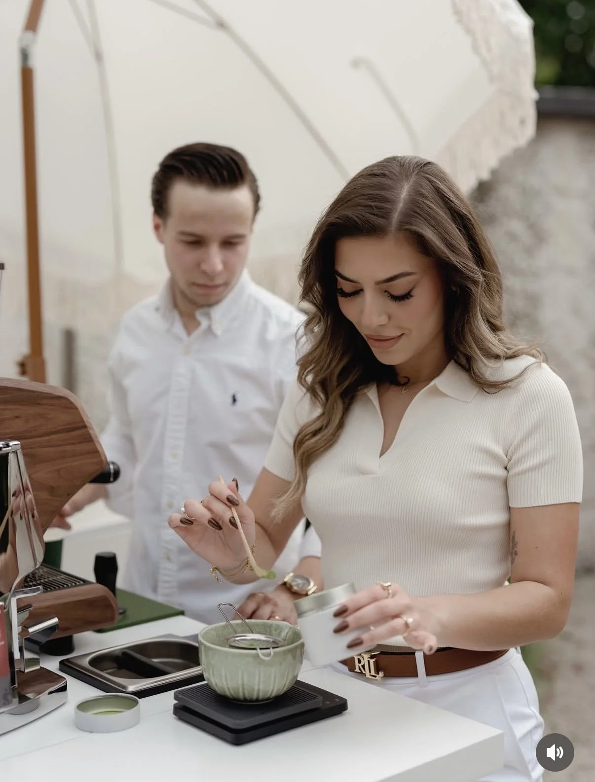 A woman and a man preparing mtcha at an outdoor coffee cart. The woman is holding a tea infuser, and the man is standing behind her. The scene includes a ceramic teacup, a portable stove, and tea-making tools.