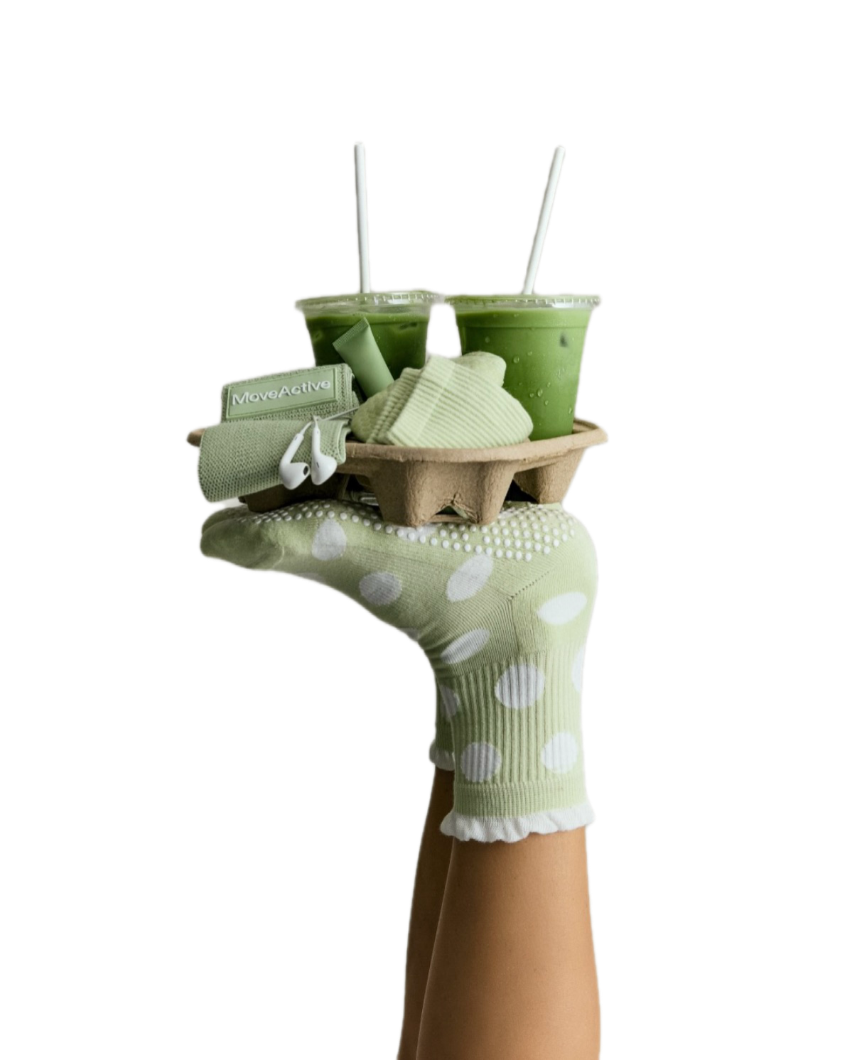 A hand wearing a green and white polka-dotted sock holds a cardboard tray with two matchas with straws, a pair of white earbuds, a small green towel, and a white face mask, against a black background.