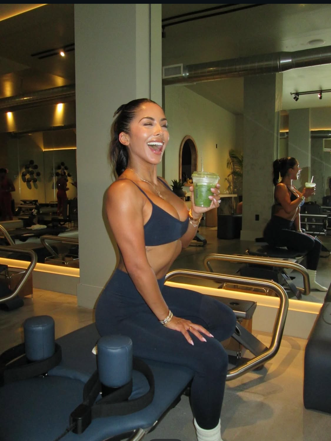 A woman in workout attire sitting on a workout bench in a gym, holding an iced matcha, smiling and laughing after pilates class.
