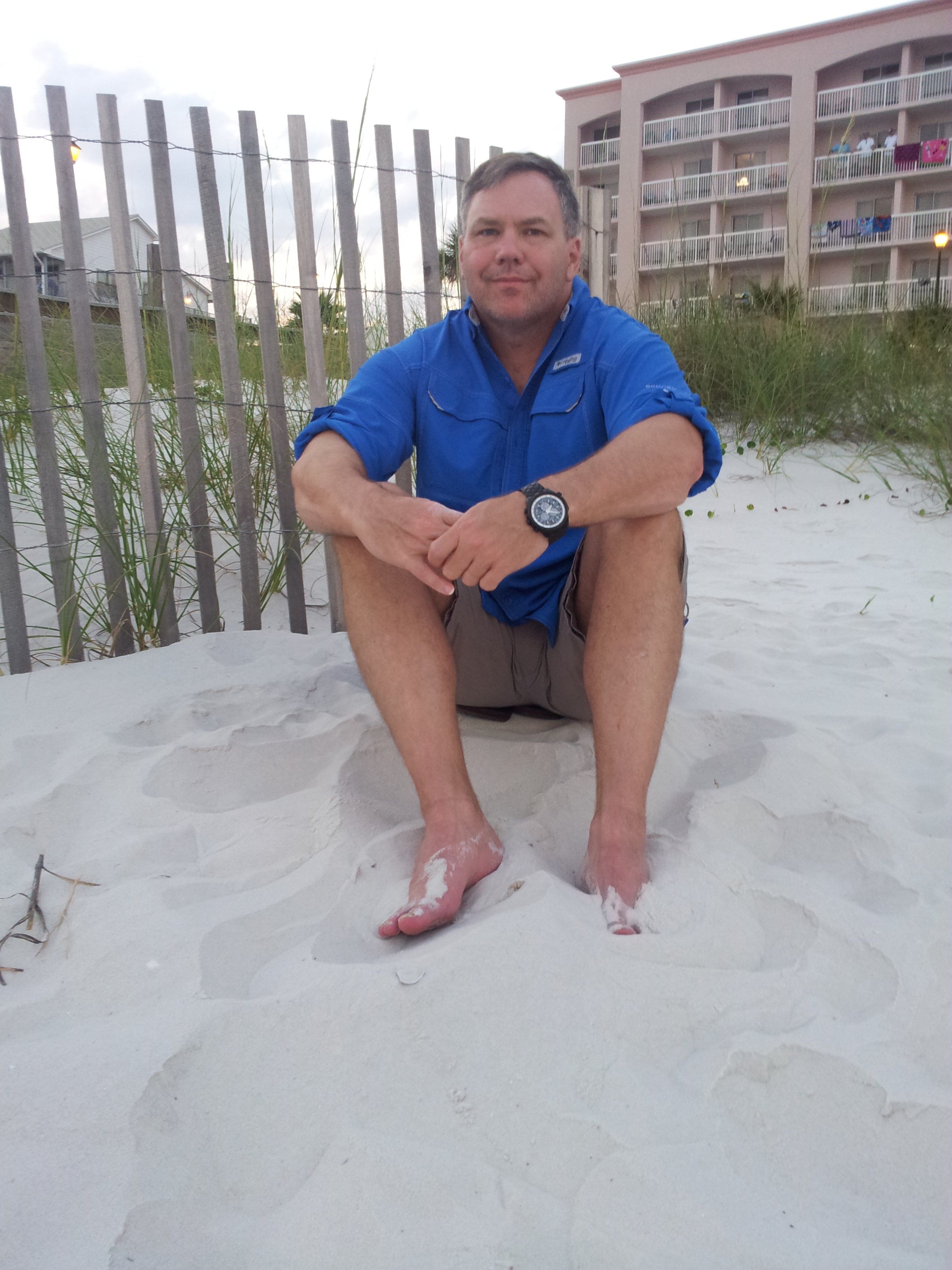 A man sitting on white sandy beach sand with his legs crossed, wearing a blue shirt, khakis, and a wristwatch, with a background of a beachside building, grass, fence, and cloudy sky.