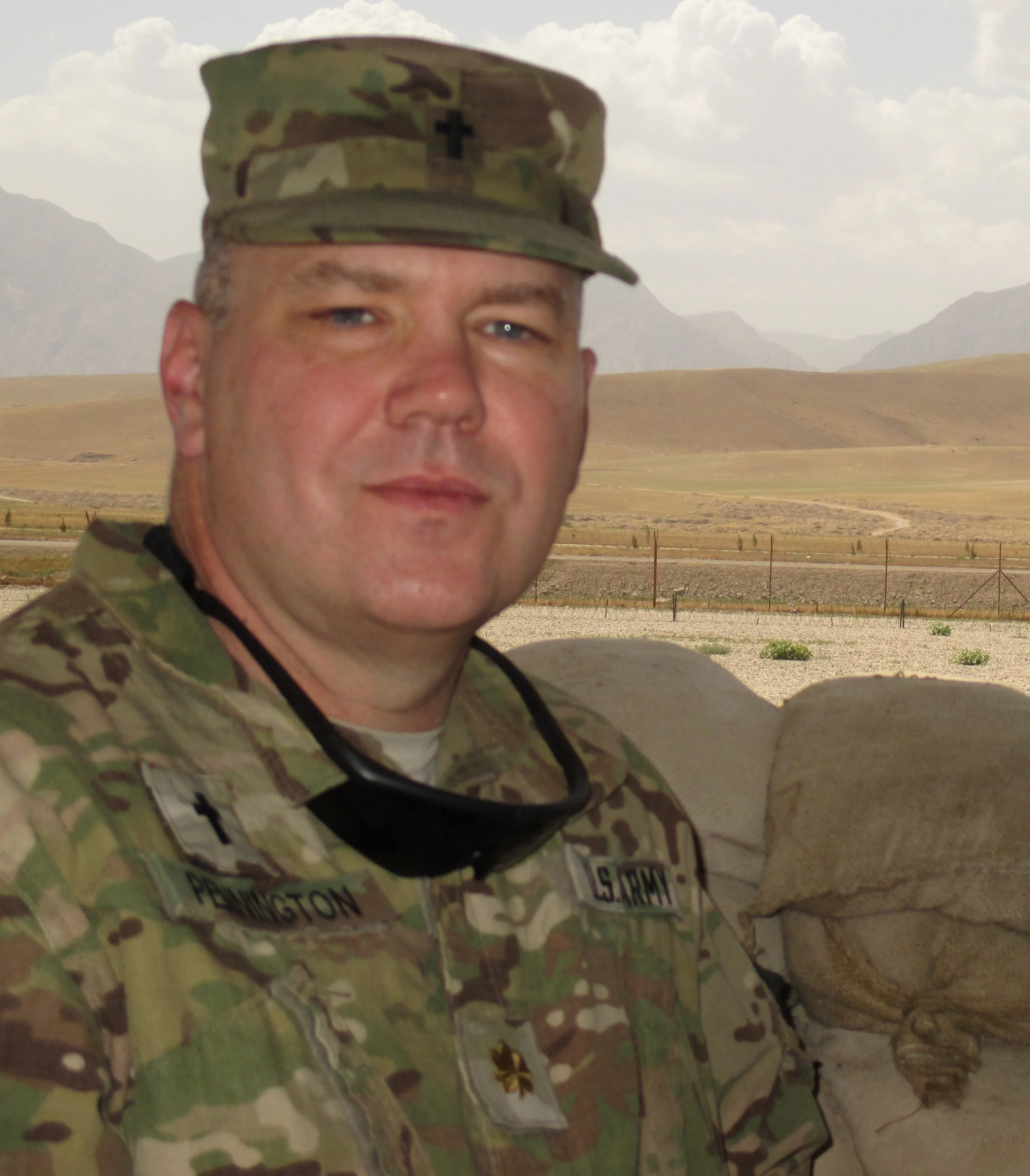 A person in camouflage military uniform with a camouflage cap, standing outdoors with mountains and cloudy sky in the background.