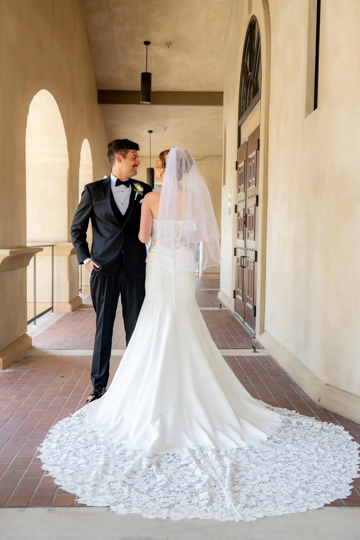 A bride and groom are standing in a hallway, gazing at each other with smiles. The bride is dressed in a white wedding gown with a train and veil, and the groom is in a black tuxedo with a bow tie and boutonniere.