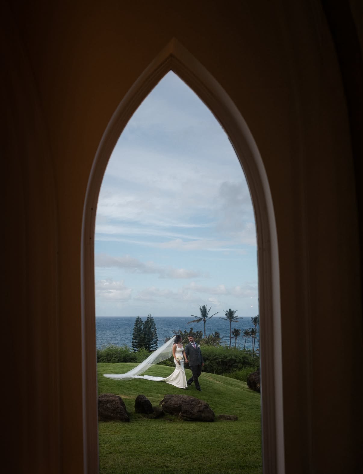 A bride and groom walking outside near the ocean, framed by a pointed arch window, with palm trees and cloudy sky in the background.