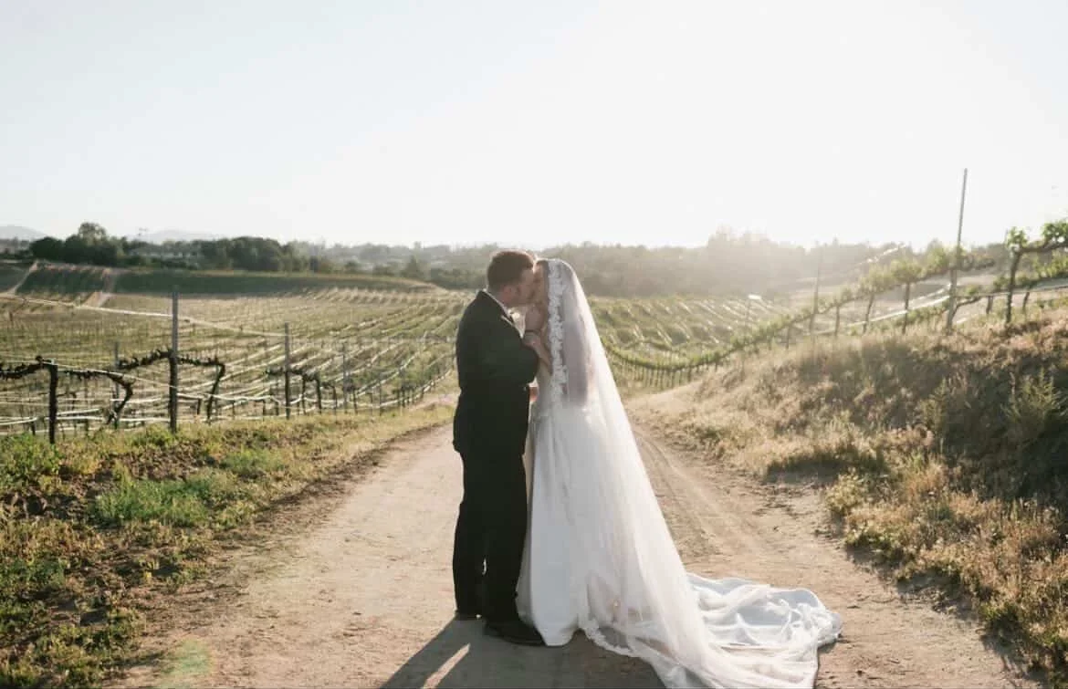 A bride and groom kissing on a dirt path in a vineyard during sunset.