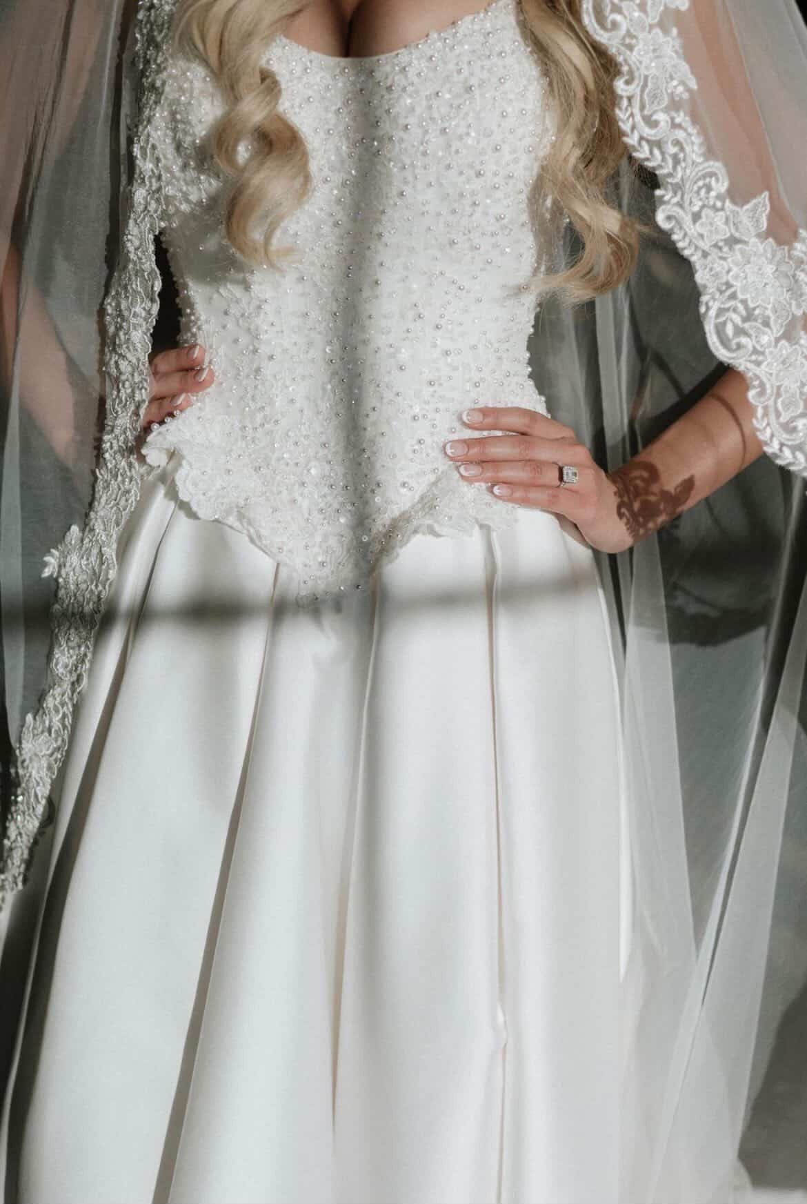 A woman in a white wedding dress with pearl embellishments and lace sleeves, standing with hands on hips, wearing a ring and a tattooed arm visible, in front of a mirror.