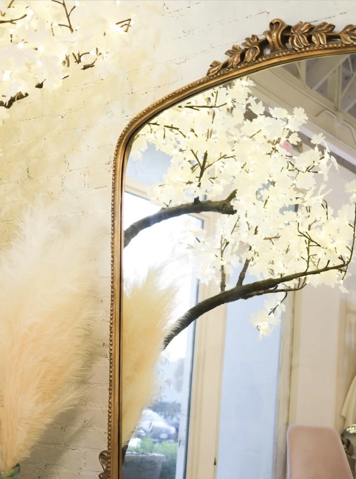 A mirror reflecting artificial white blossoms on a tree branch, with a yellow-colored wall, pampas grass, and part of a pink chair visible in the room.