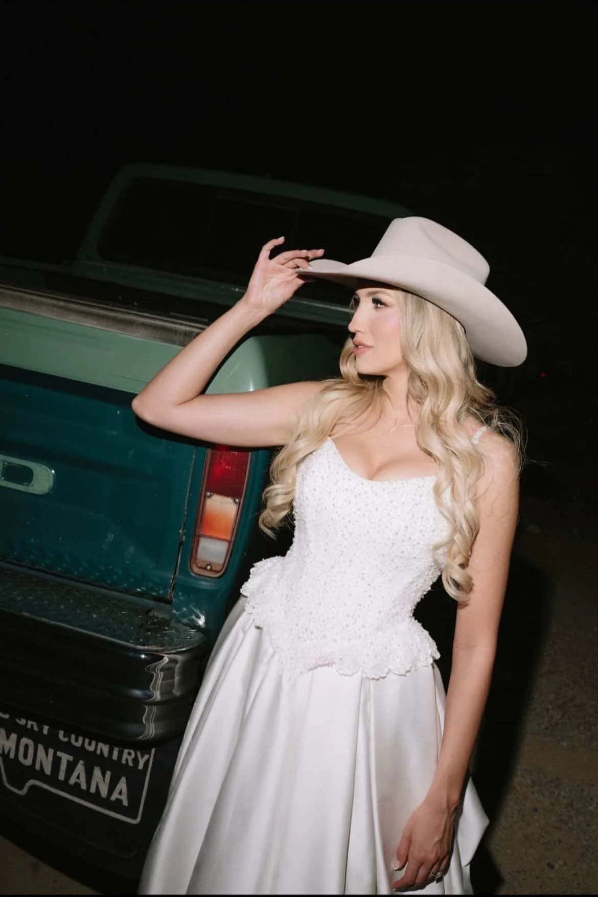 A woman with long blonde hair wearing a white dress and a large white cowboy hat, standing next to a green truck at night.