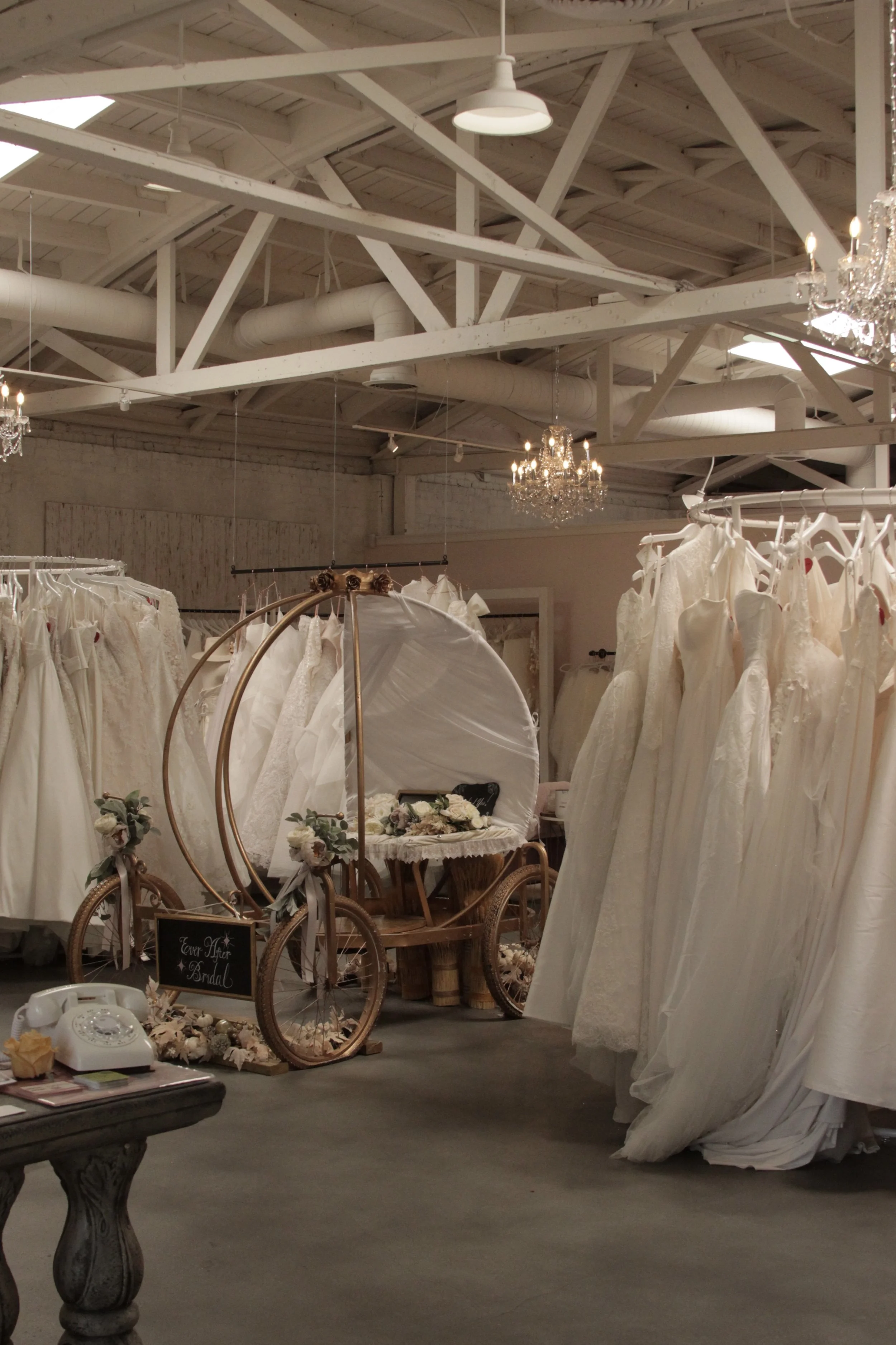 A wedding dress boutique with numerous white wedding gowns on racks and a vintage-style bicycle decorated with flowers and a blackboard sign for a wedding after-party, set inside a spacious industrial-style room with white exposed beams and chandeliers.