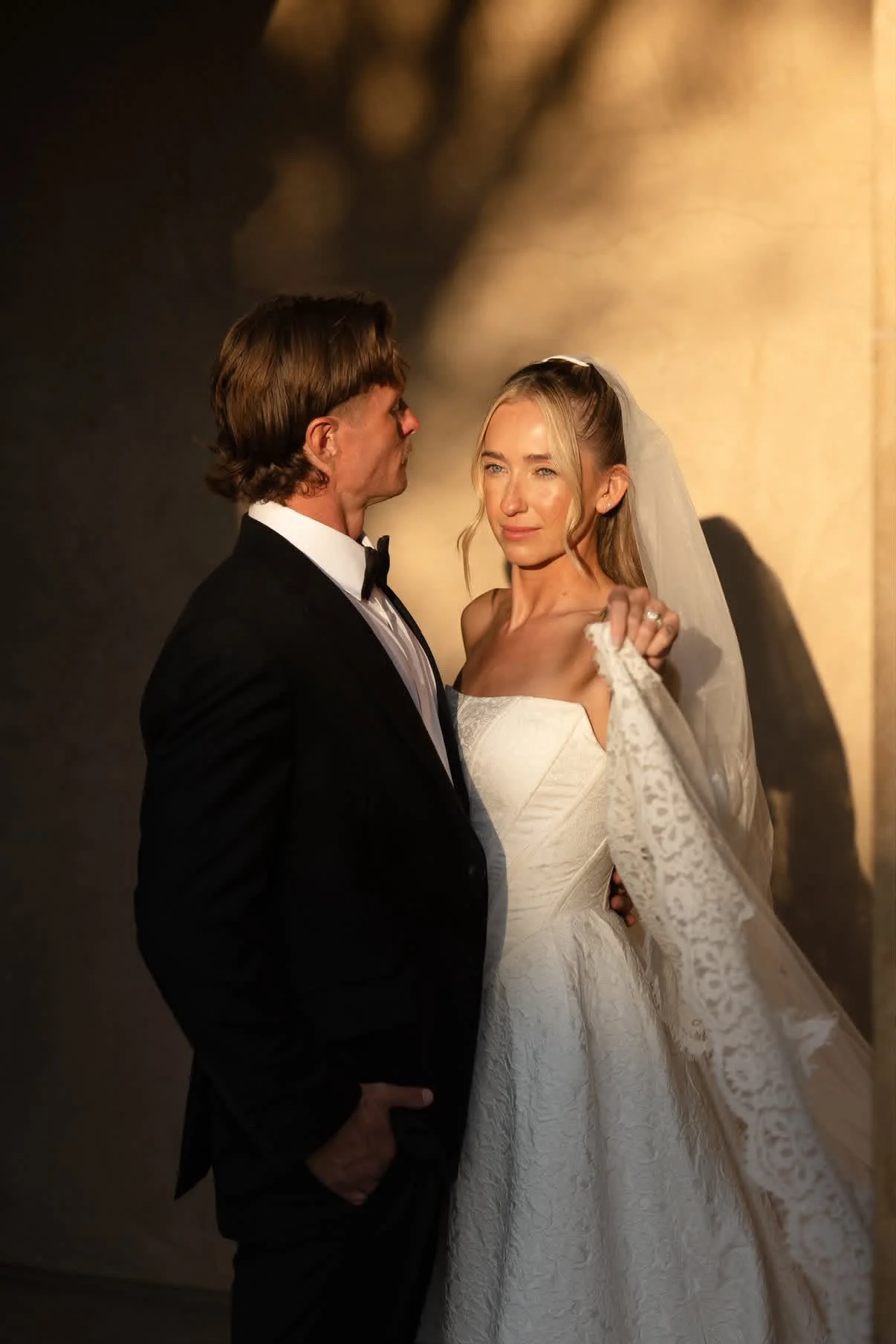 A bride in a white wedding dress and veil standing close to a groom in a black tuxedo, during their wedding photoshoot. The bride is holding her veil with her right hand, and they are indoors with a backdrop of warm lighting and shadows.