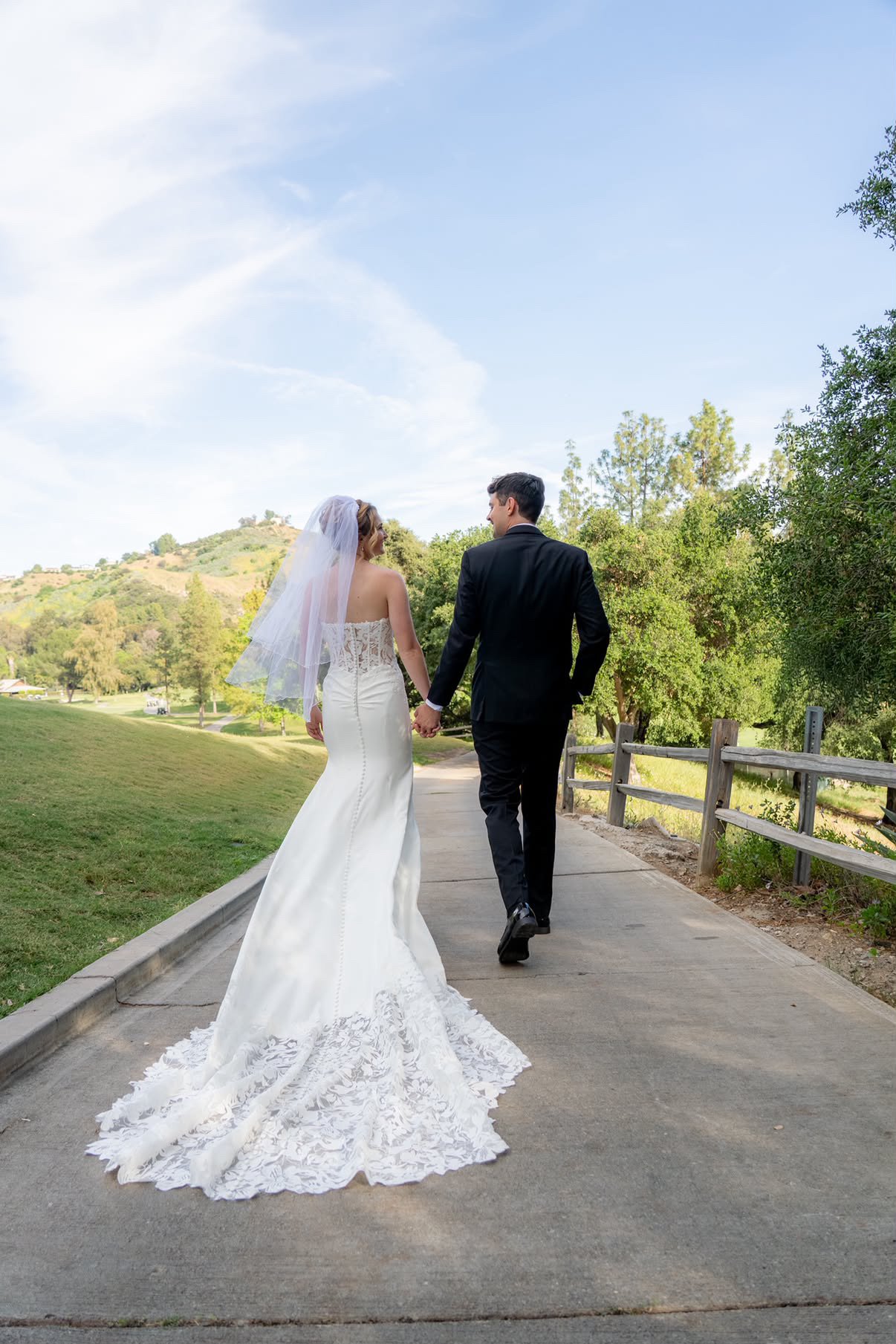 A bride and groom walk hand in hand on a park trail with trees and a hill in the background, the bride wearing a white wedding dress and veil, and the groom in a black suit.