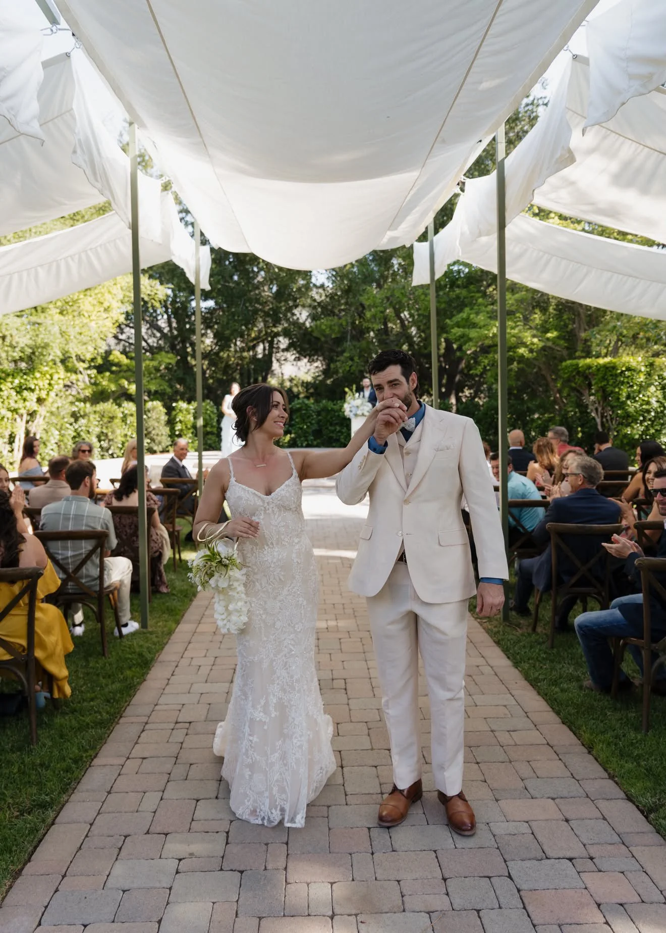 A bride and groom walking down an outdoor aisle surrounded by seated wedding guests, with a canopy of white fabric overhead and lush green trees in the background.